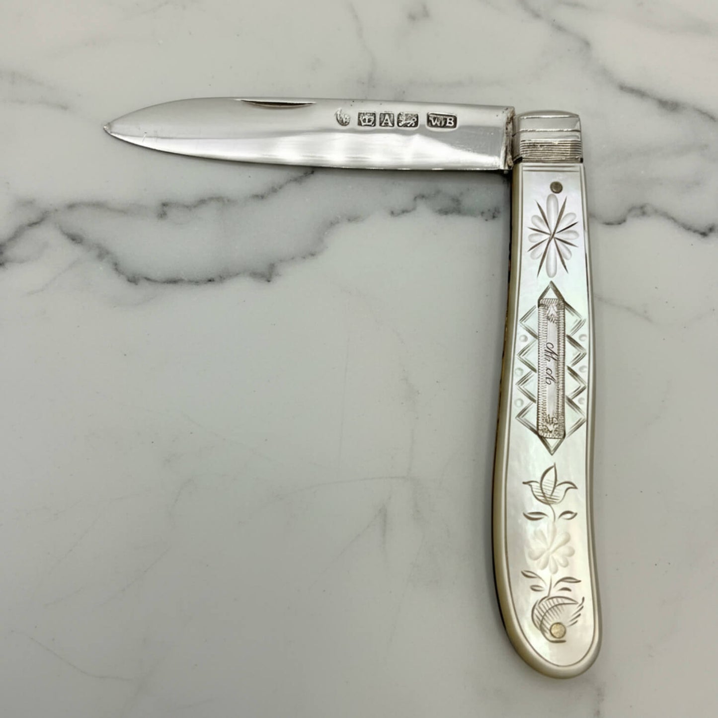 A vintage sterling silver folding pocket knife with engraved floral and geometric patterns on its mother of pearl handle, displayed open on a white marble surface.