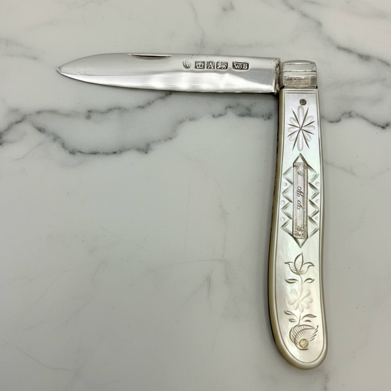 A vintage sterling silver folding pocket knife with engraved floral and geometric patterns on its mother of pearl handle, displayed open on a white marble surface.