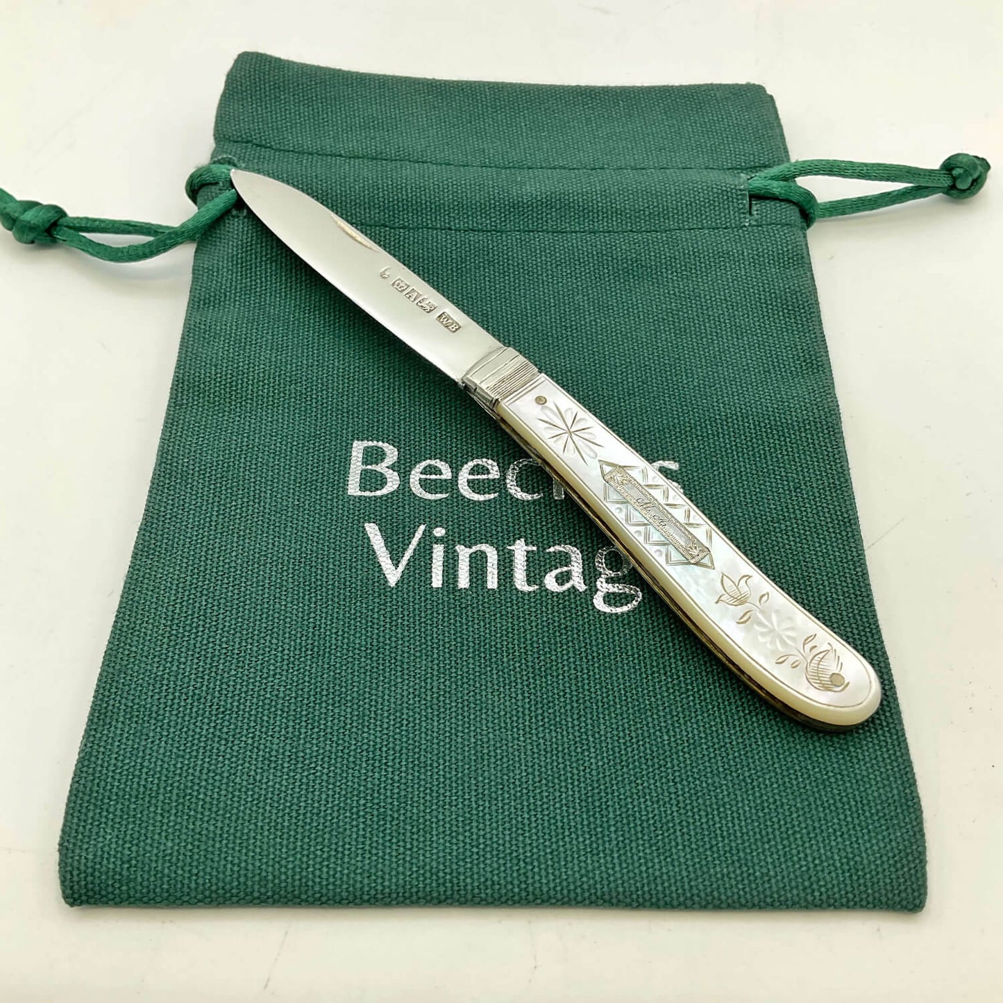 An antique silver fruit knife with an engraved handle, resting diagonally on a dark green canvas drawstring pouch with 'Beeches Vintage' printed on it.