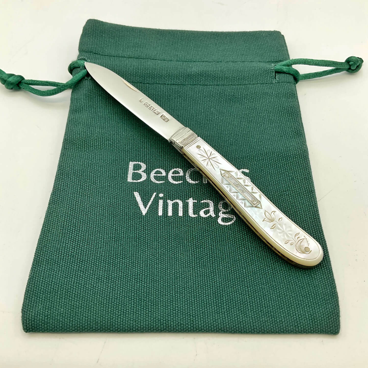 An antique silver fruit knife with an engraved handle, resting diagonally on a dark green canvas drawstring pouch with 'Beeches Vintage' printed on it.