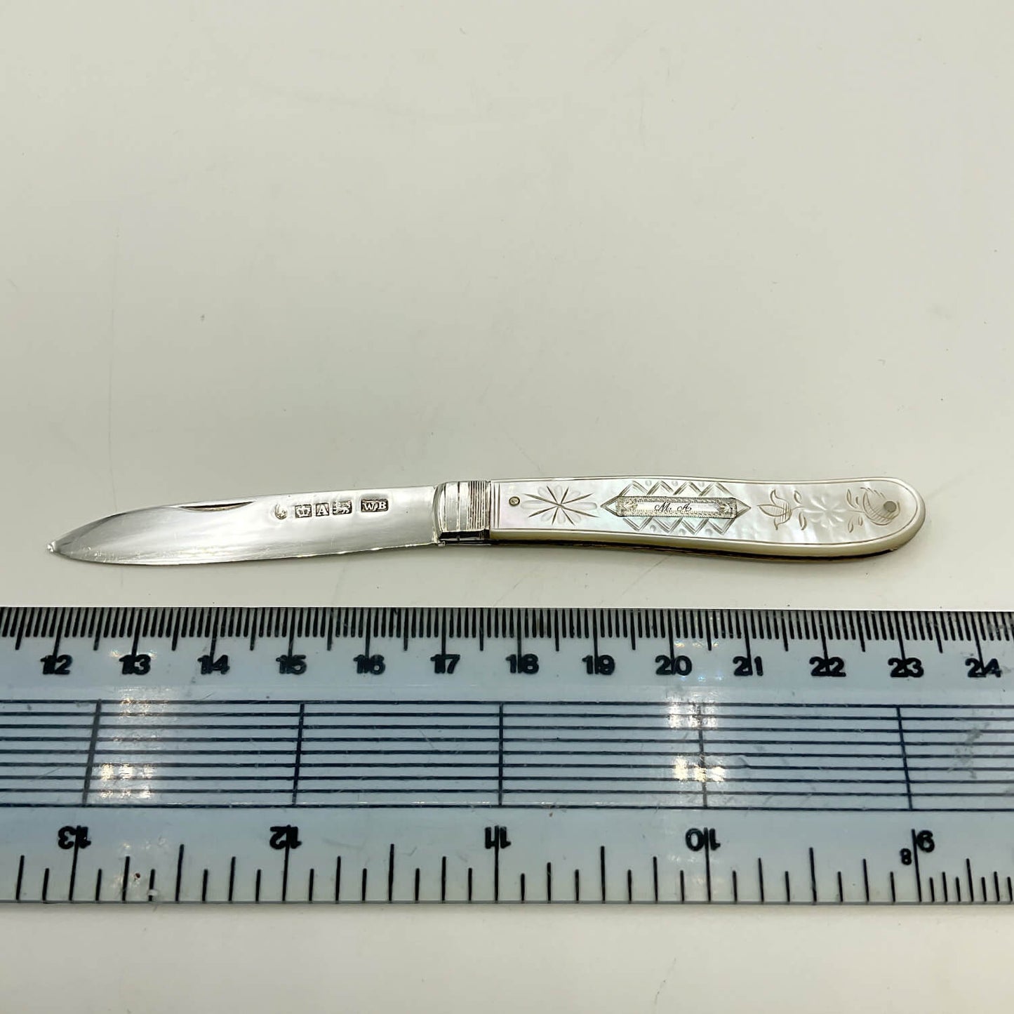 A vintage sterling silver pocket knife with engraved floral and geometric patterns on its handle, displayed horizontally above a clear plastic ruler for scale on a light off-white background.