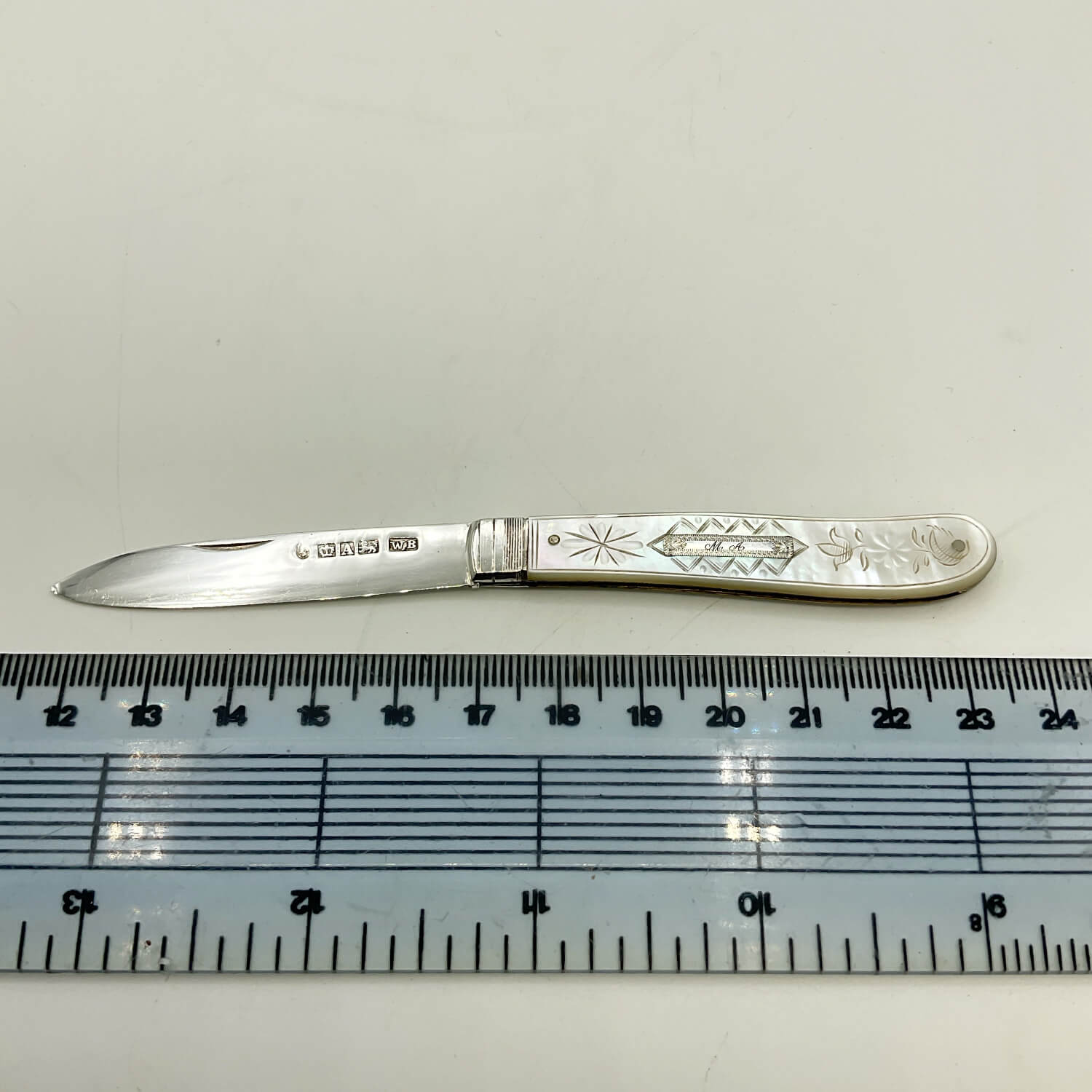 A vintage sterling silver pocket knife with engraved floral and geometric patterns on its handle, displayed horizontally above a clear plastic ruler for scale on a light off-white background.