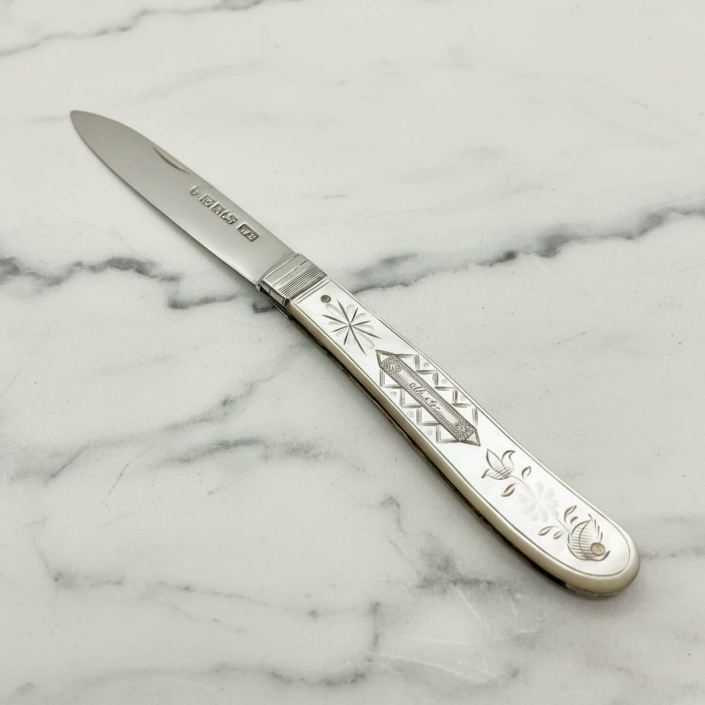 An antique silver knife with a silver blade and an intricately engraved MOP handle featuring mother-of-pearl floral details, resting on a light gray surface.