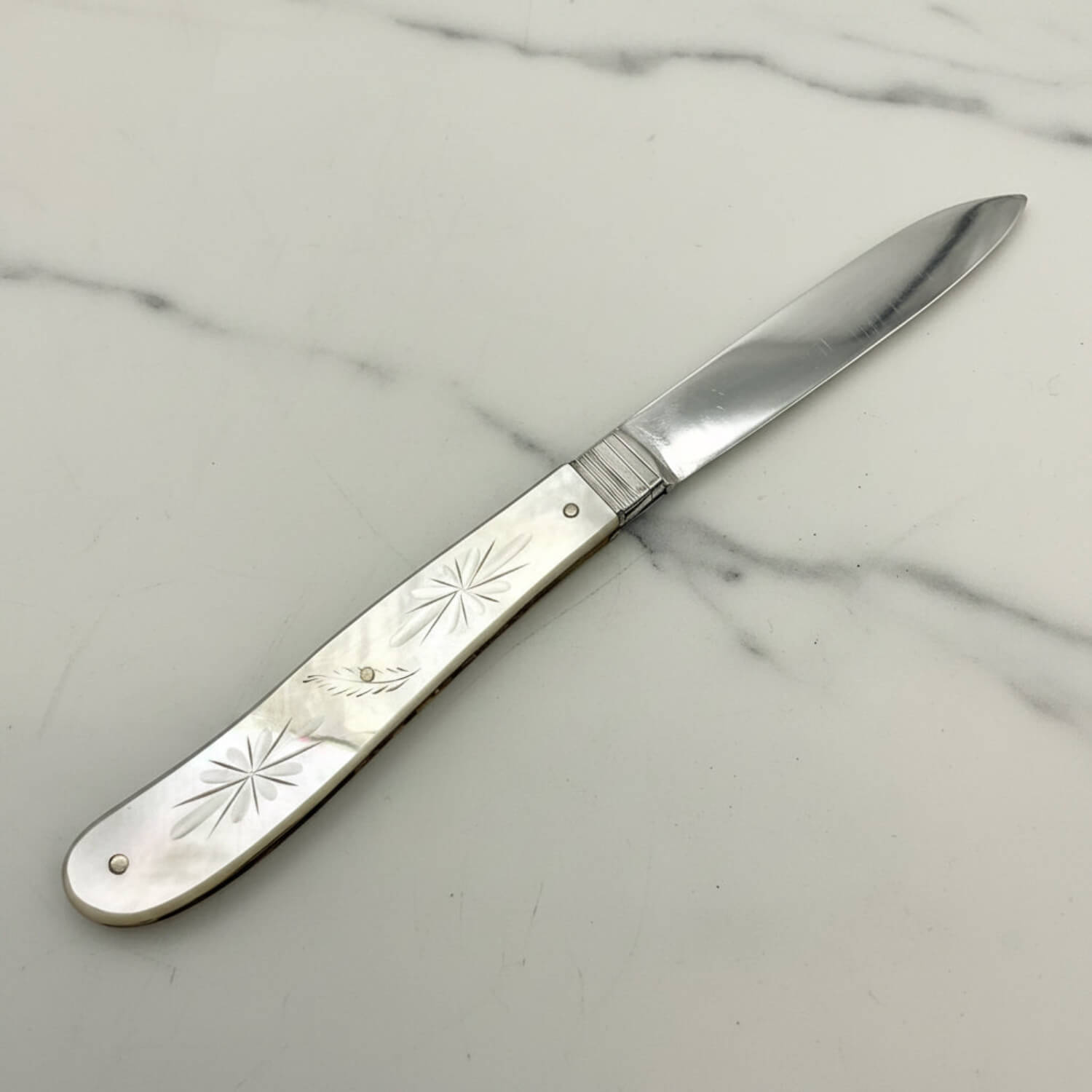 AAntique silver knife with an engraved mother-of-pearl handle and a polished silver blade, shown on a neutral light gray background.