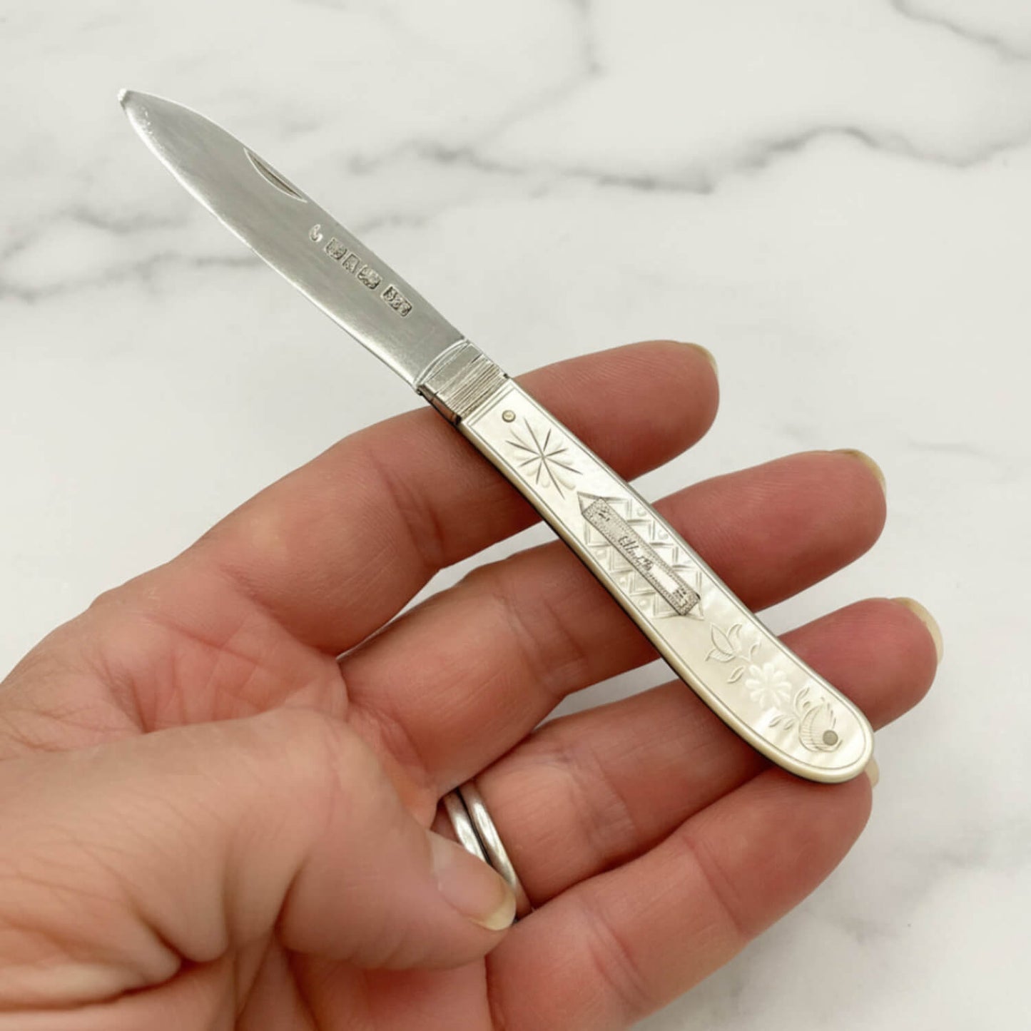 A vintage engraved pocket knife with a silver blade and a decorative mother of pearl handle, held in a person's hand against a plain white background.