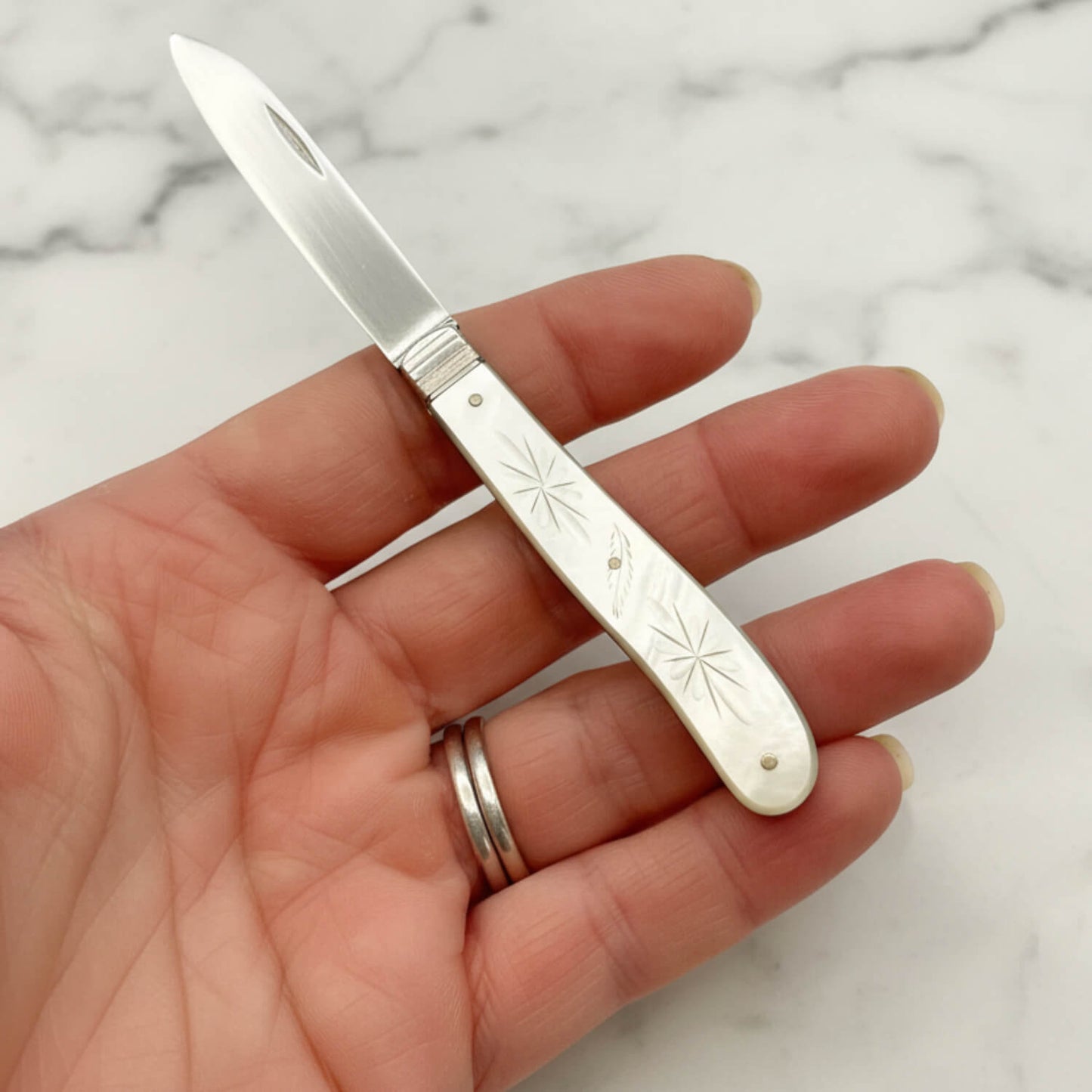 A silver pocket knife with an engraved white handle, held in a person's hand against a white marble background.