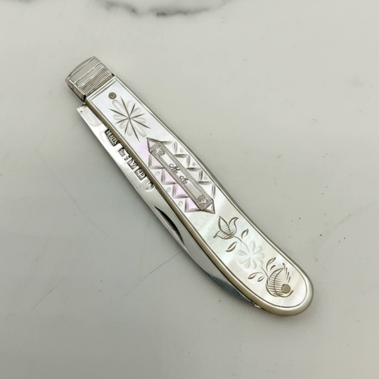 A vintage pocket knife with a silver handle featuring mother-of-pearl floral inlays and engraved details, shown open on a light marble surface.
