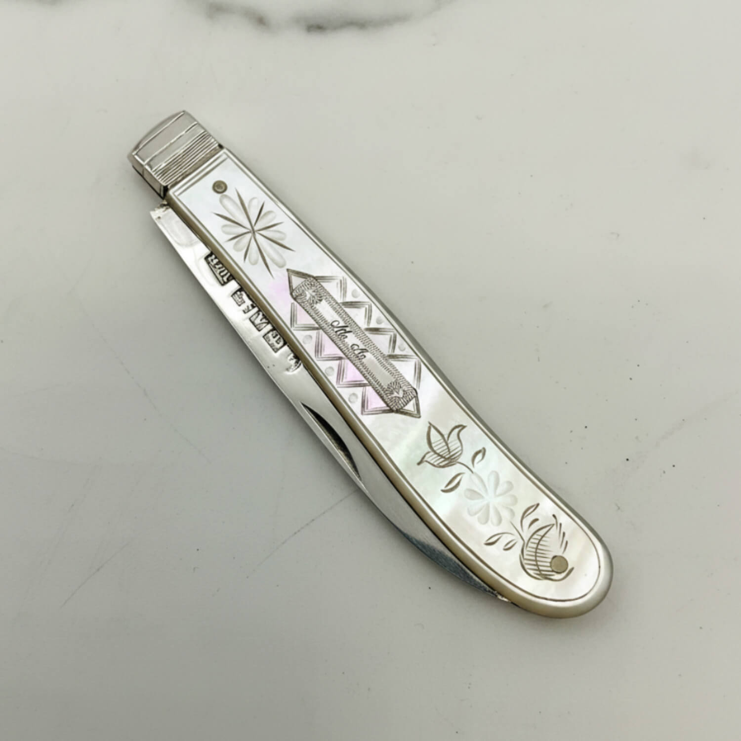 A vintage pocket knife with a silver handle featuring mother-of-pearl floral inlays and engraved details, shown open on a light marble surface.