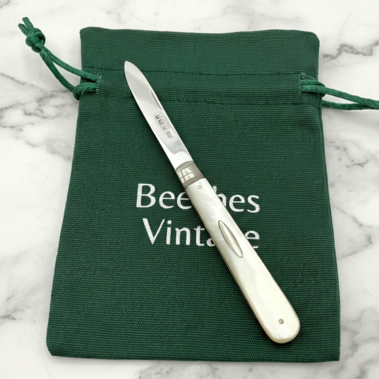 Silver fruit knife with white handle on a green drawstring bag labeled 'Beeches Vintage' on a marble surface.