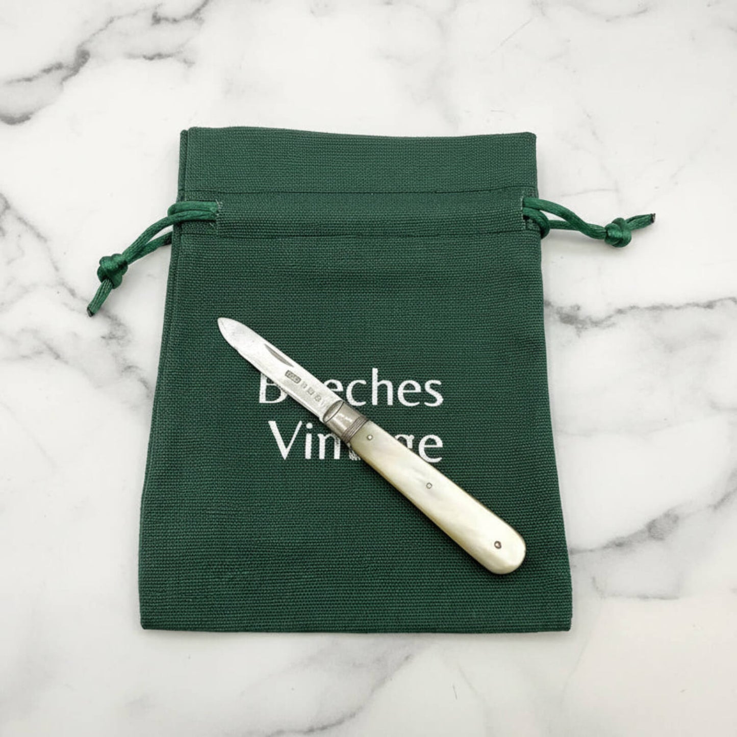 Knife with white handle on a green drawstring bag labeled 'Beeches Vintage' on a marble background