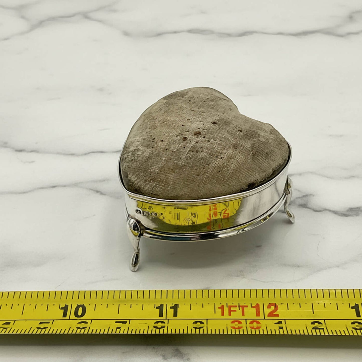 Heart-shaped pincushion on a silver stand with a measuring tape for scale on a marble surface