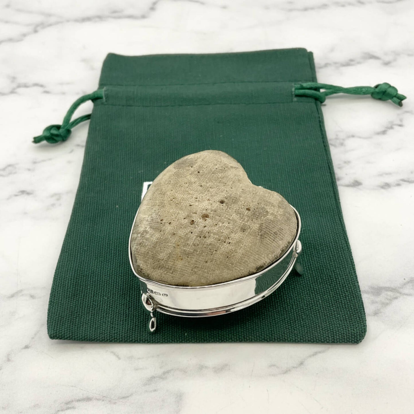 Heart-shaped pincushion in a silver holder on a green drawstring bag with a marble background