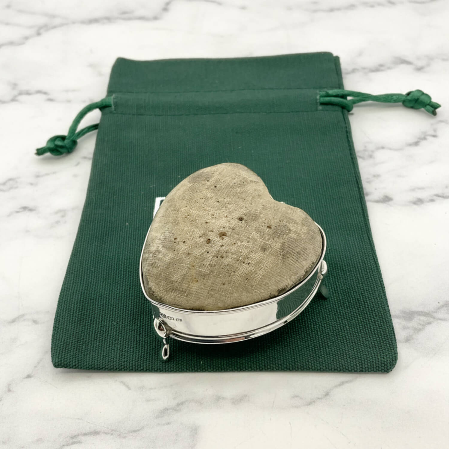 Heart-shaped pincushion in a silver holder on a green drawstring bag with a marble background