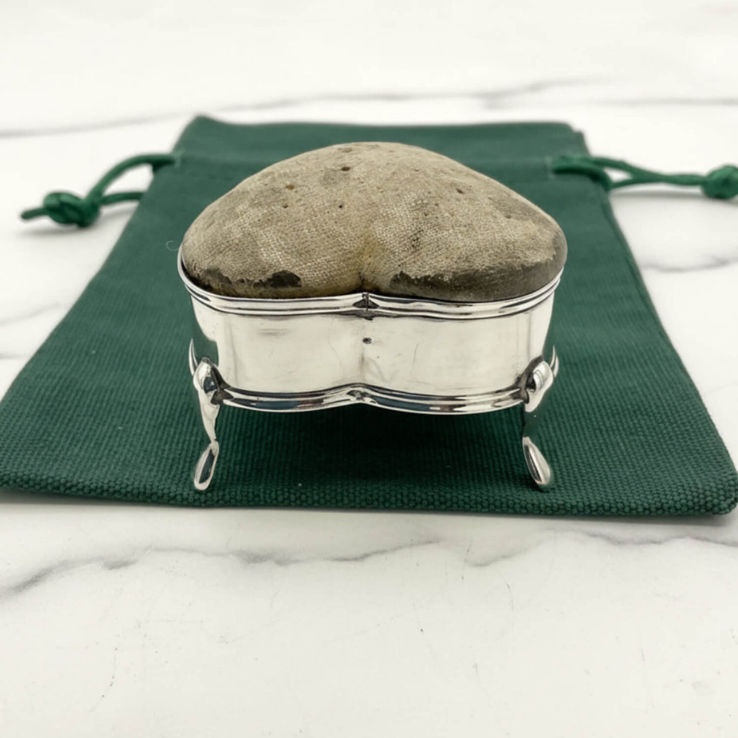 Heart-shaped pincushion in a silver holder on a green cloth with a white marble background