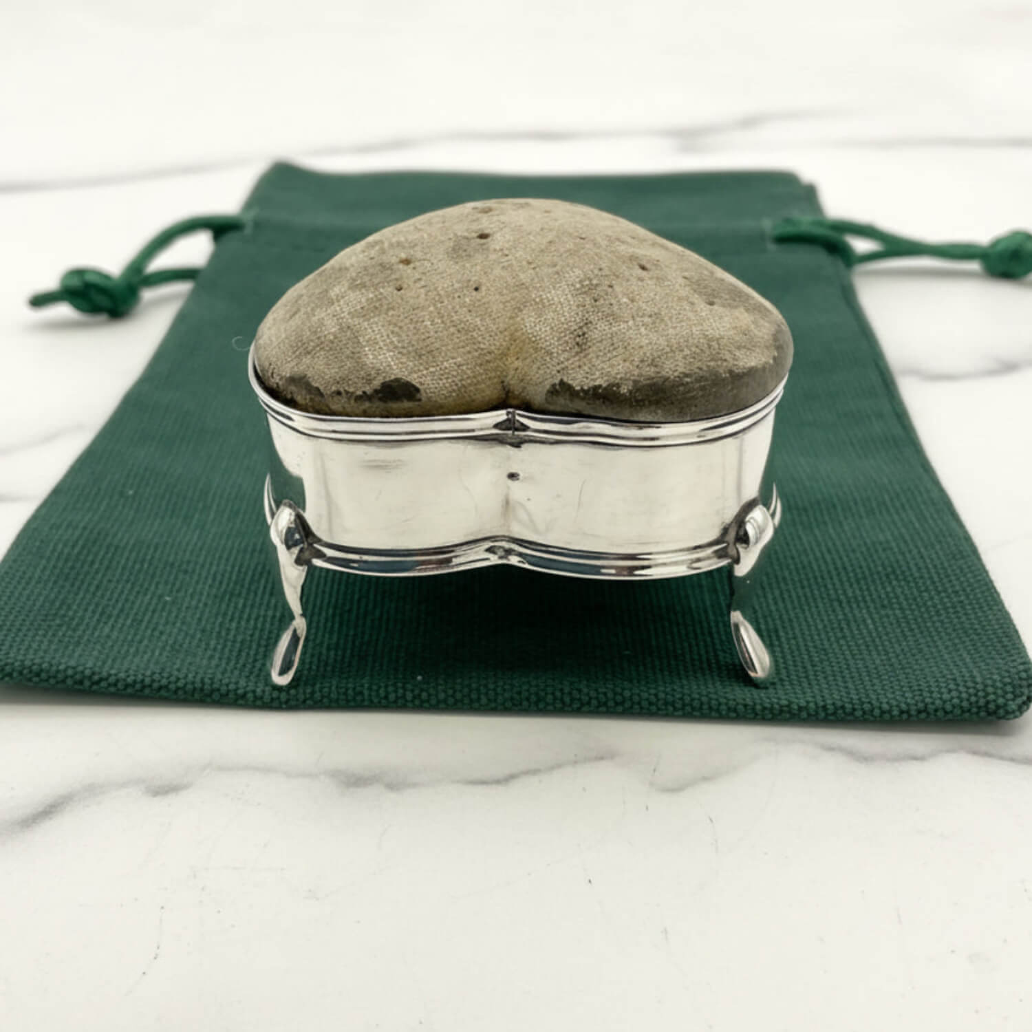 Heart-shaped pincushion in a silver holder on a green cloth with a white marble background