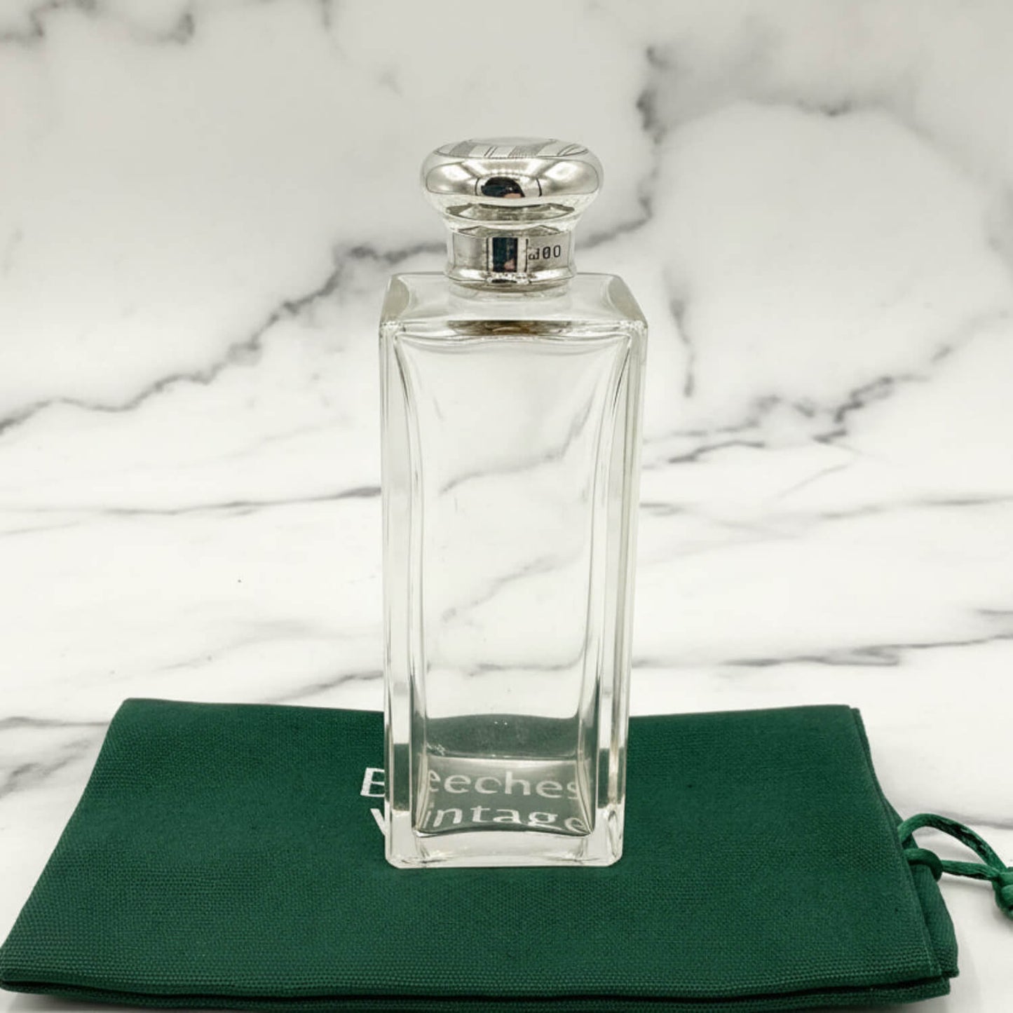 Clear glass perfume bottle with silver cap on a green cloth with 'Beeches Vintage' branding against a marble background.