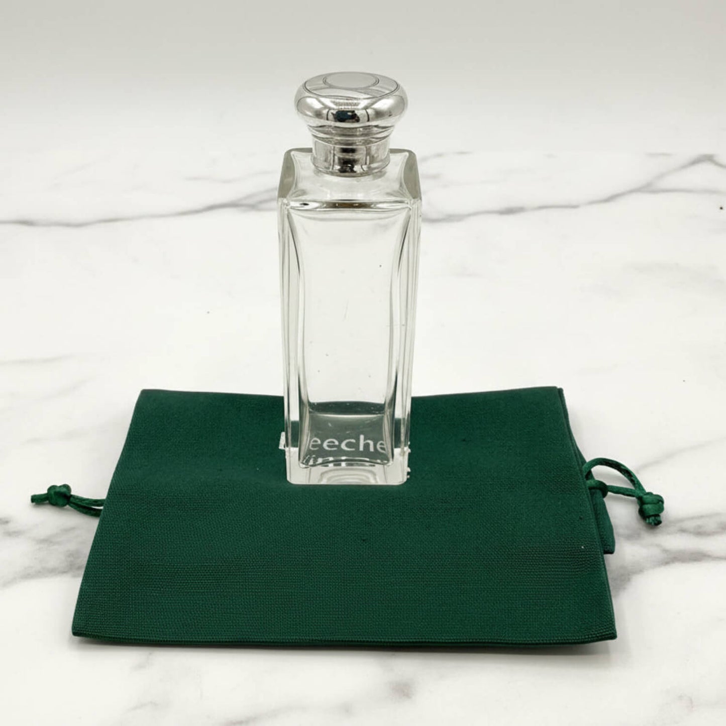 Clear glass bottle with silver cap on a green drawstring bag against a marble background