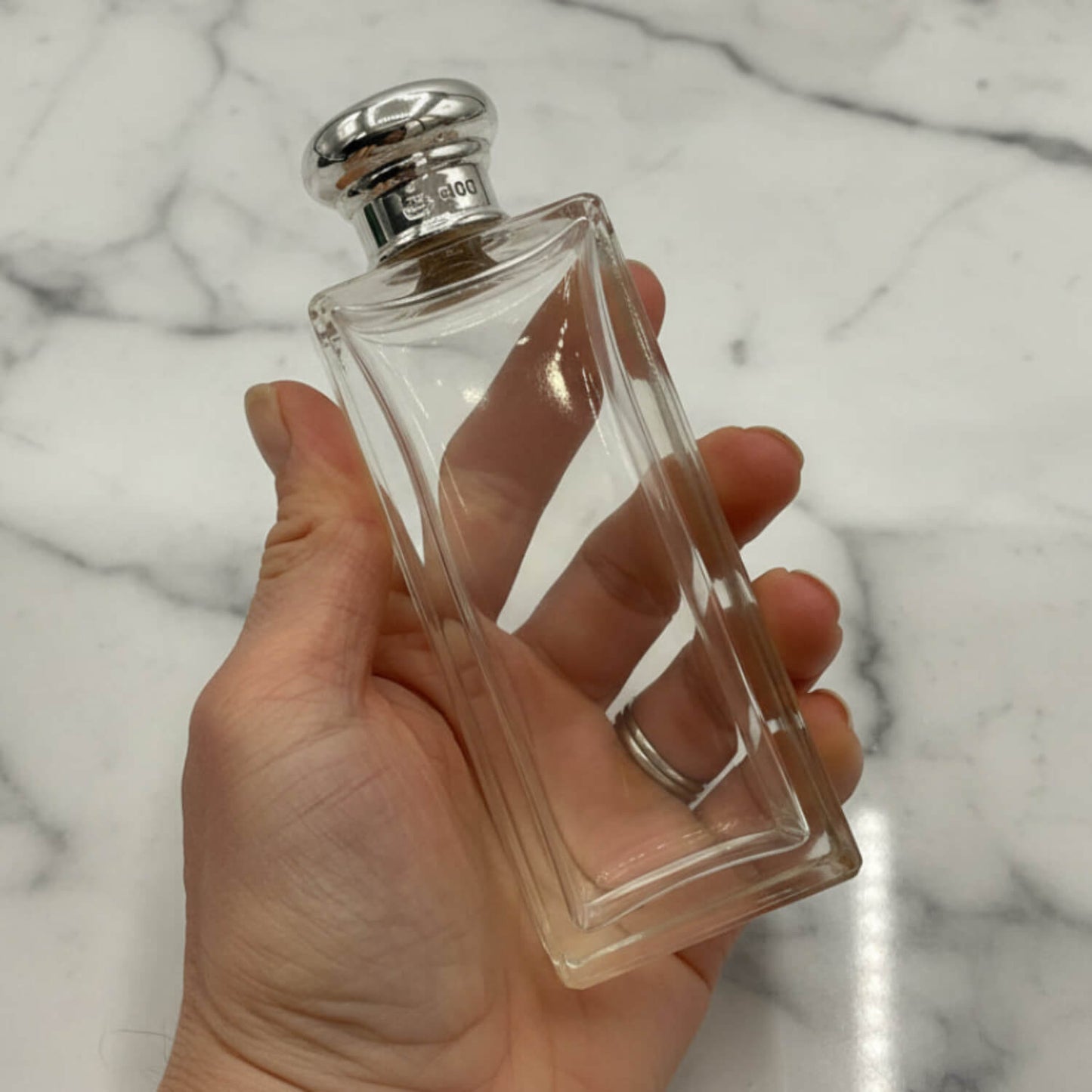 Clear perfume bottle with silver cap held in a hand against a marble background