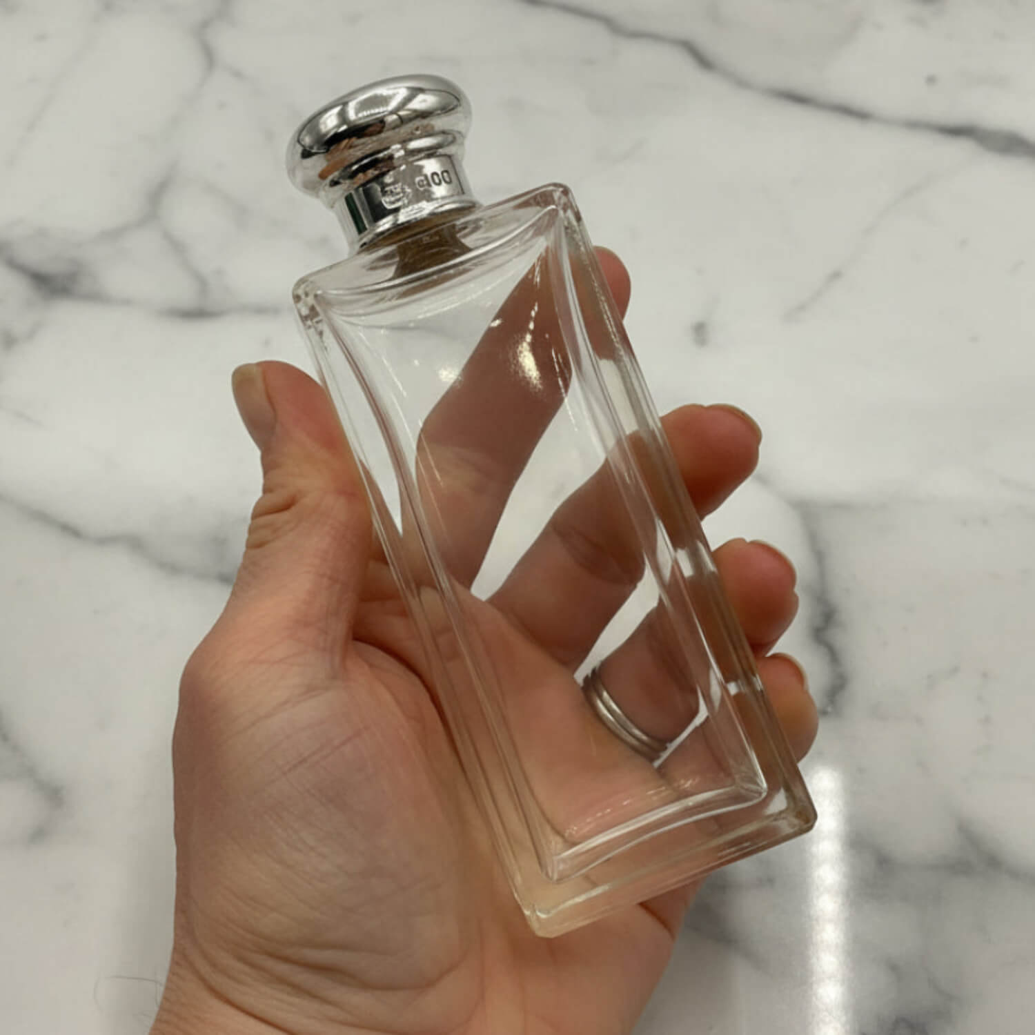 Clear perfume bottle with silver cap held in a hand against a marble background