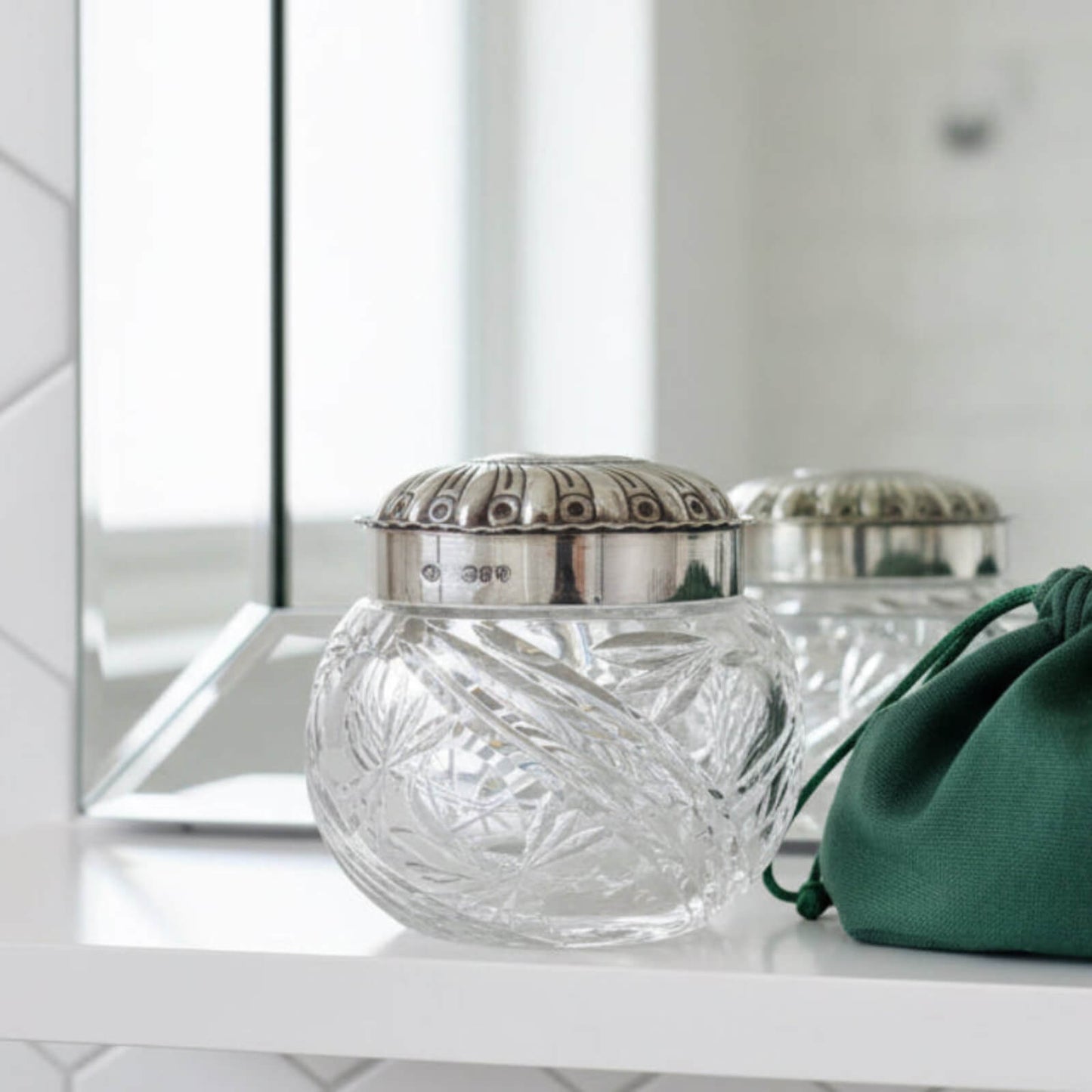 Decorative glass jar with silver lid on a white surface, with a green pouch in the foreground.