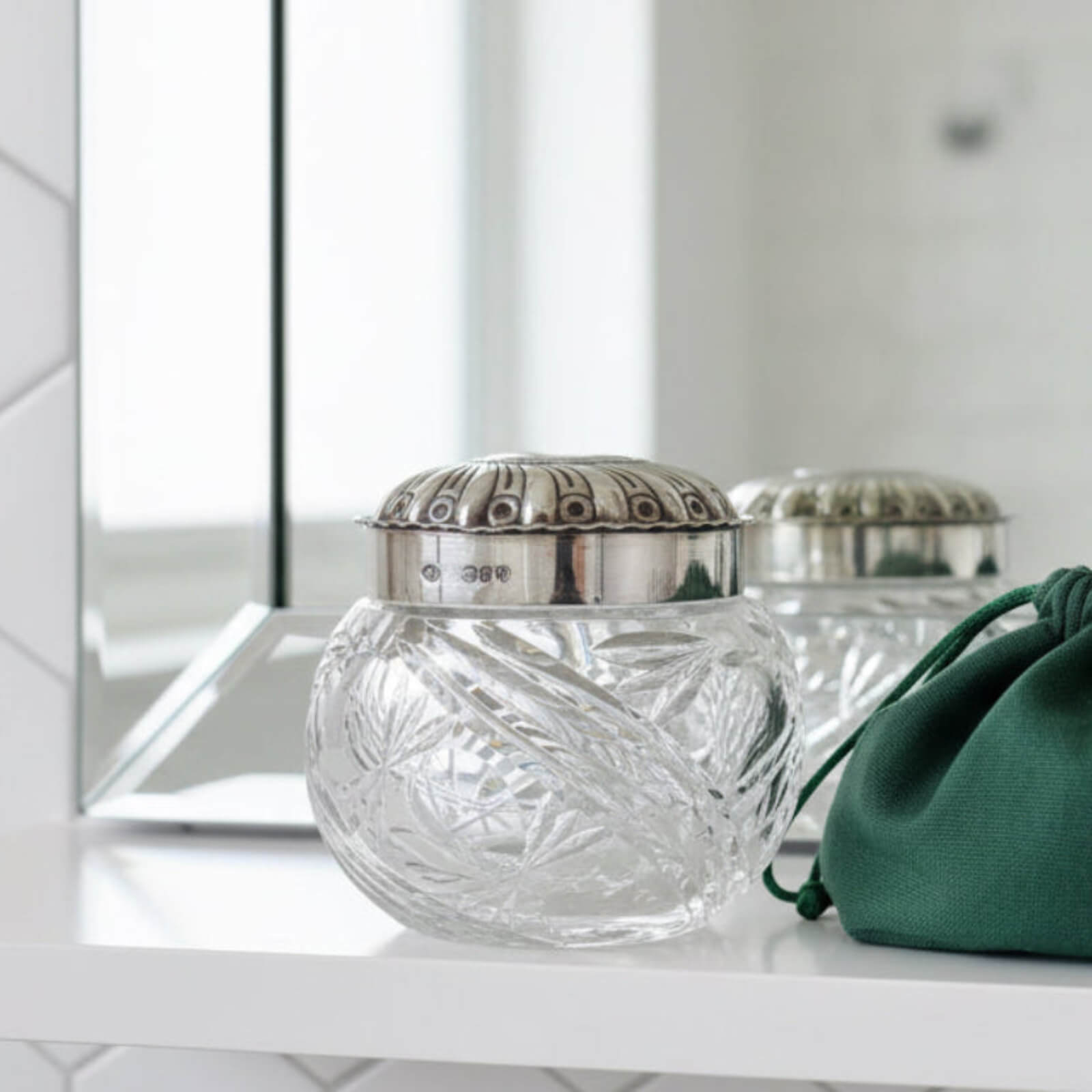 Decorative glass jar with silver lid on a white surface, with a green pouch in the foreground.
