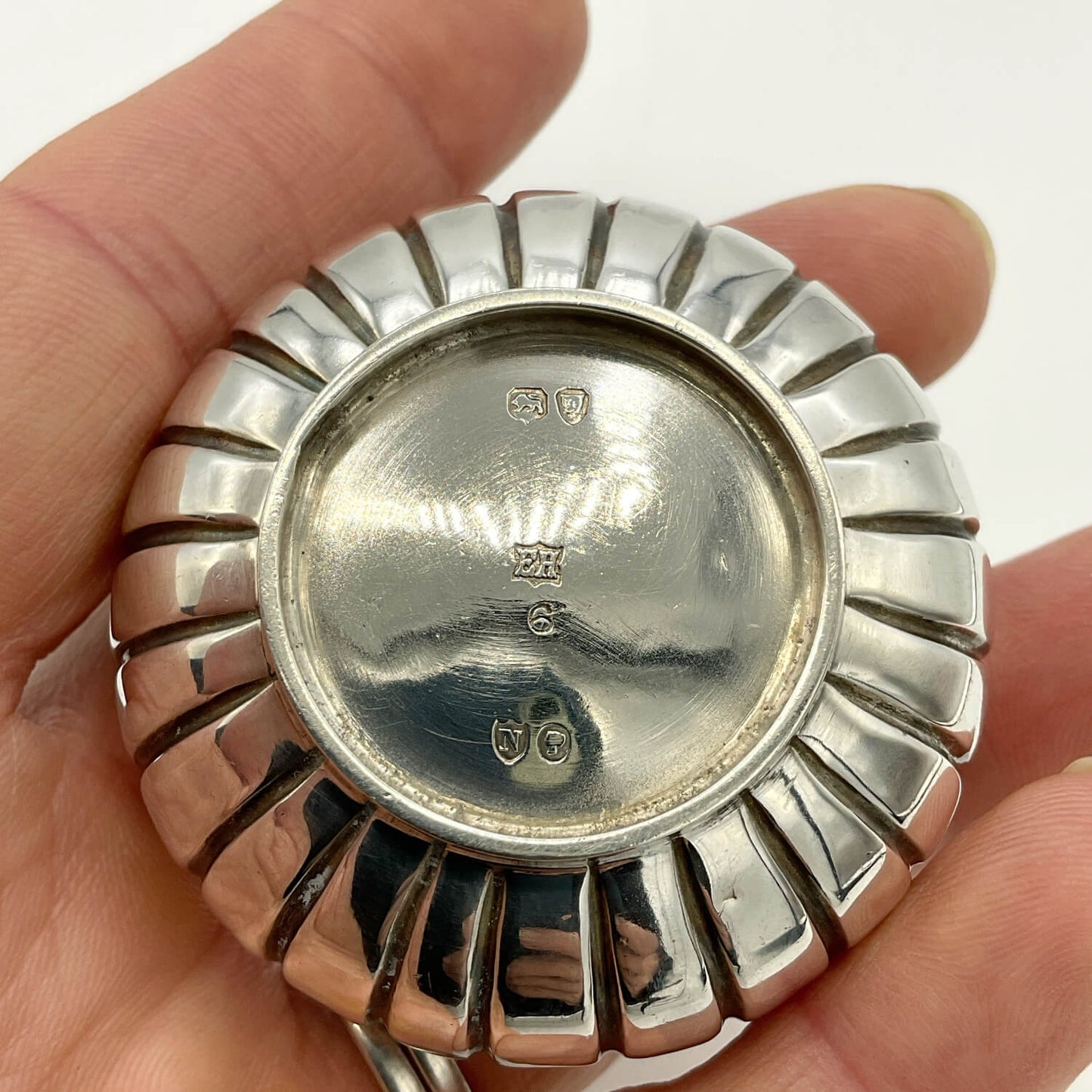 A close-up of a polished silver round box held in a hand, showing a sunburst-style segmented outer rim and engraved hallmarks on the flat top.