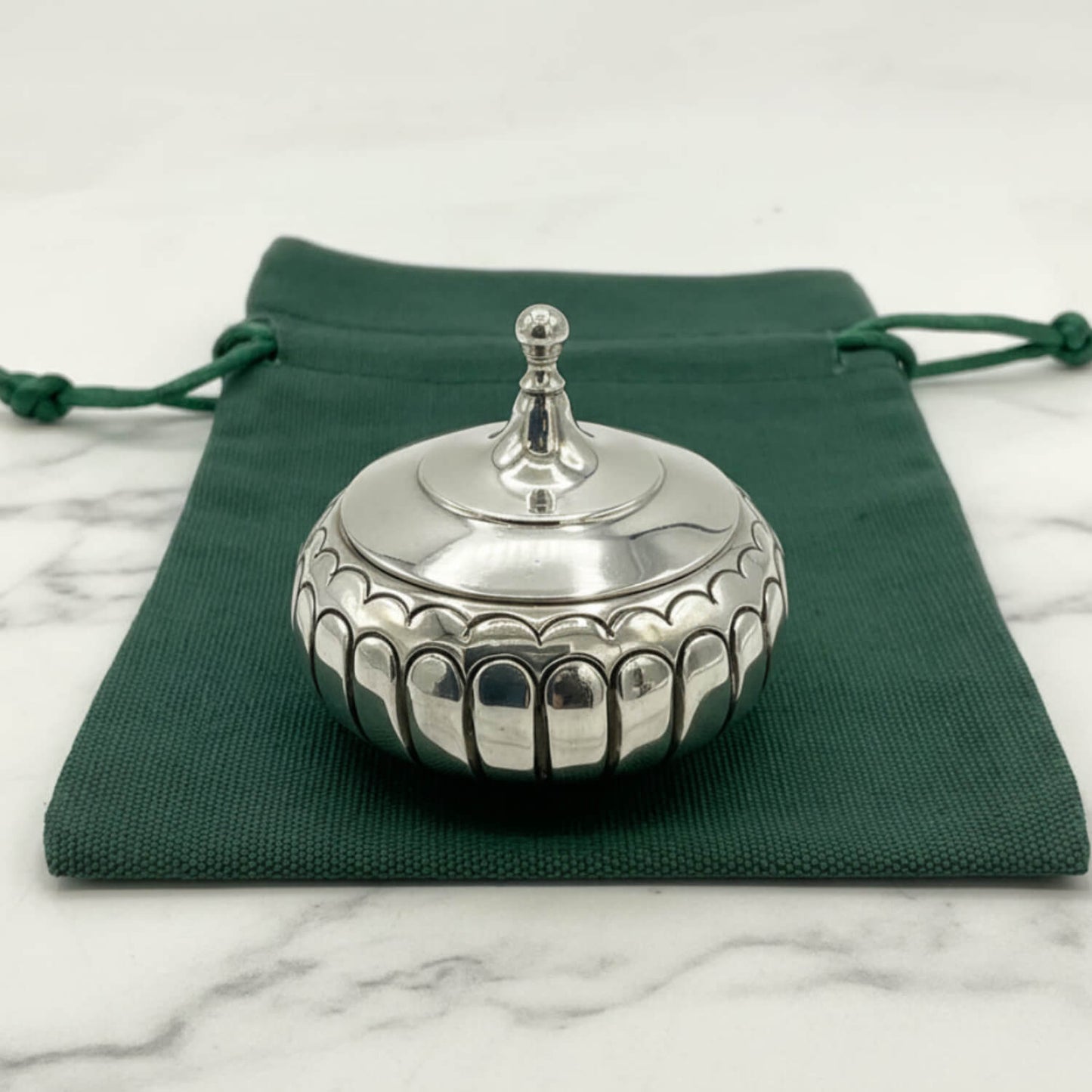 A small round silver box with a fitted lid and decorative scalloped edge, placed on top of a dark green drawstring pouch over a light marble surface.