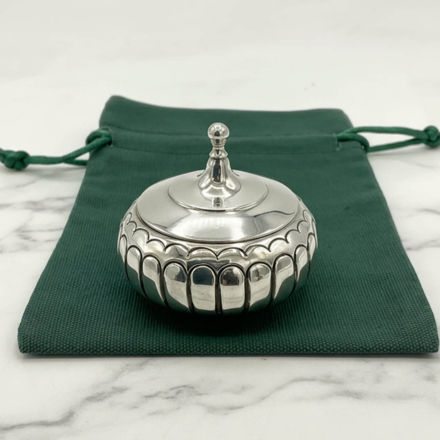 A small round silver box with a fitted lid and decorative scalloped edge, placed on top of a dark green drawstring pouch over a light marble surface.