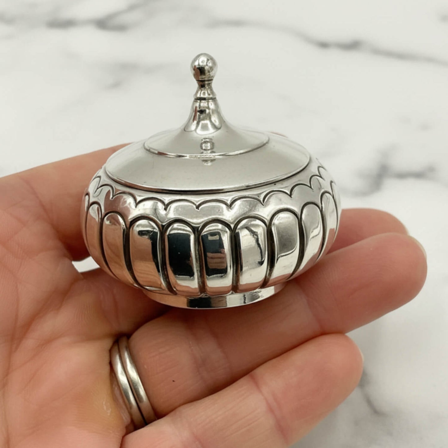 A small polished silver decorative lidded box with a pointed finial top, held in a person's hand against a light marble background.