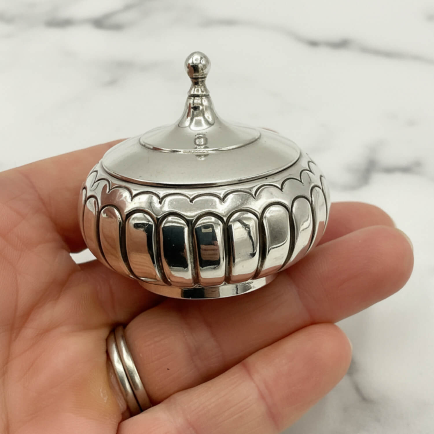 A small polished silver decorative lidded box with a pointed finial top, held in a person's hand against a light marble background.