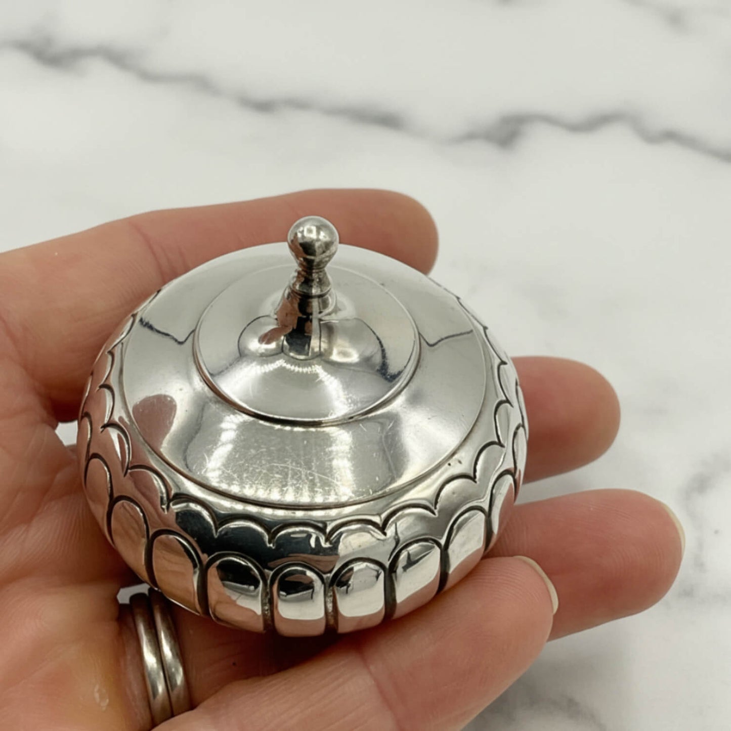 A polished silver box with a fitted lid and a small finial top, held in a person's hand against a light marble surface.