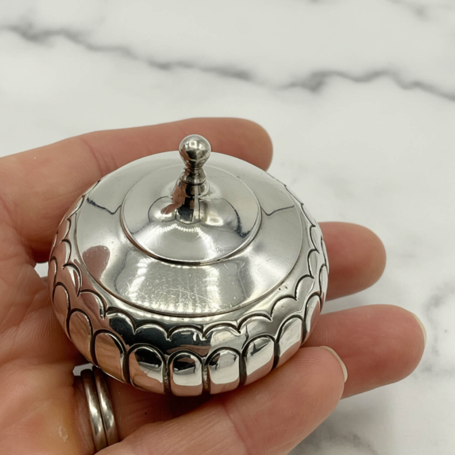 A polished silver box with a fitted lid and a small finial top, held in a person's hand against a light marble surface.