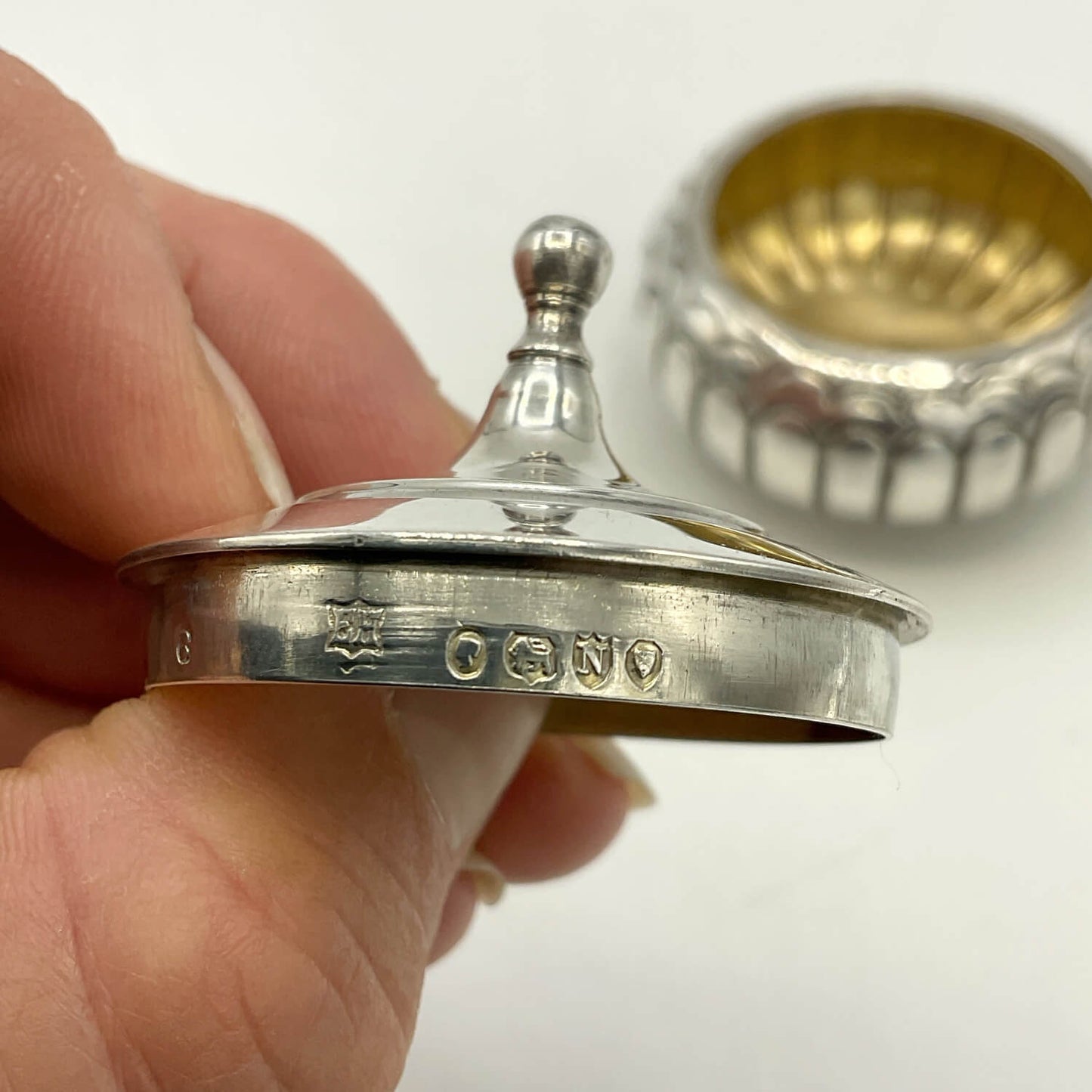 A sterling silver lidded box with a lid featuring a small finial knob, held by a hand to show the engraved hallmarks, with the gold-toned interior visible in the background.