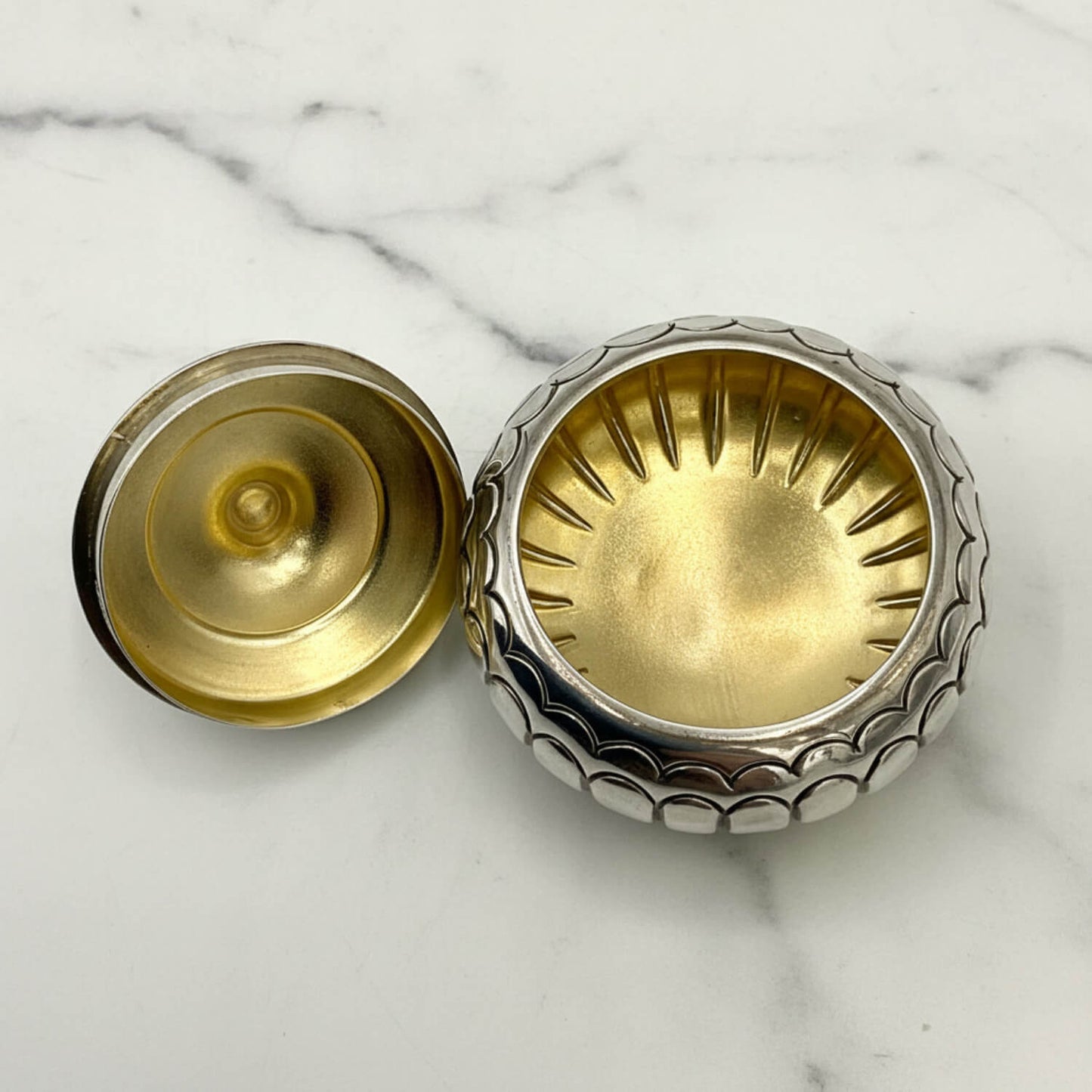 A small round silver box shown open on a white marble surface, with a silver-toned textured outer rim and a gold-toned interior and lid.