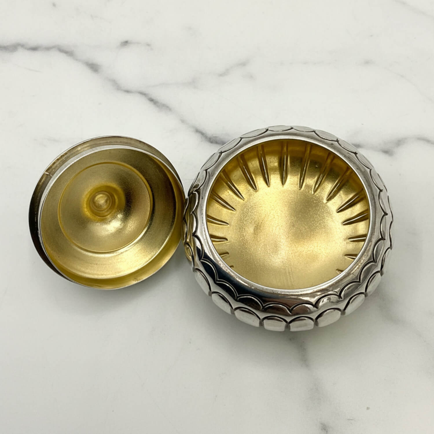 A small round silver box shown open on a white marble surface, with a silver-toned textured outer rim and a gold-toned interior and lid.