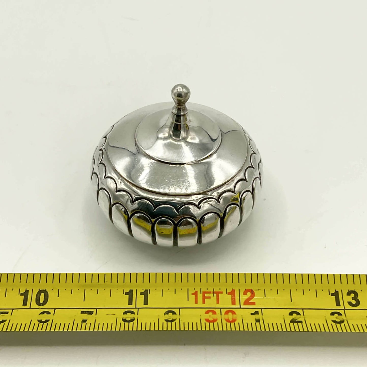 A small round silver box with a lid and decorative scalloped edge, shown on a white background with a yellow measuring tape ruler at the bottom for scale.