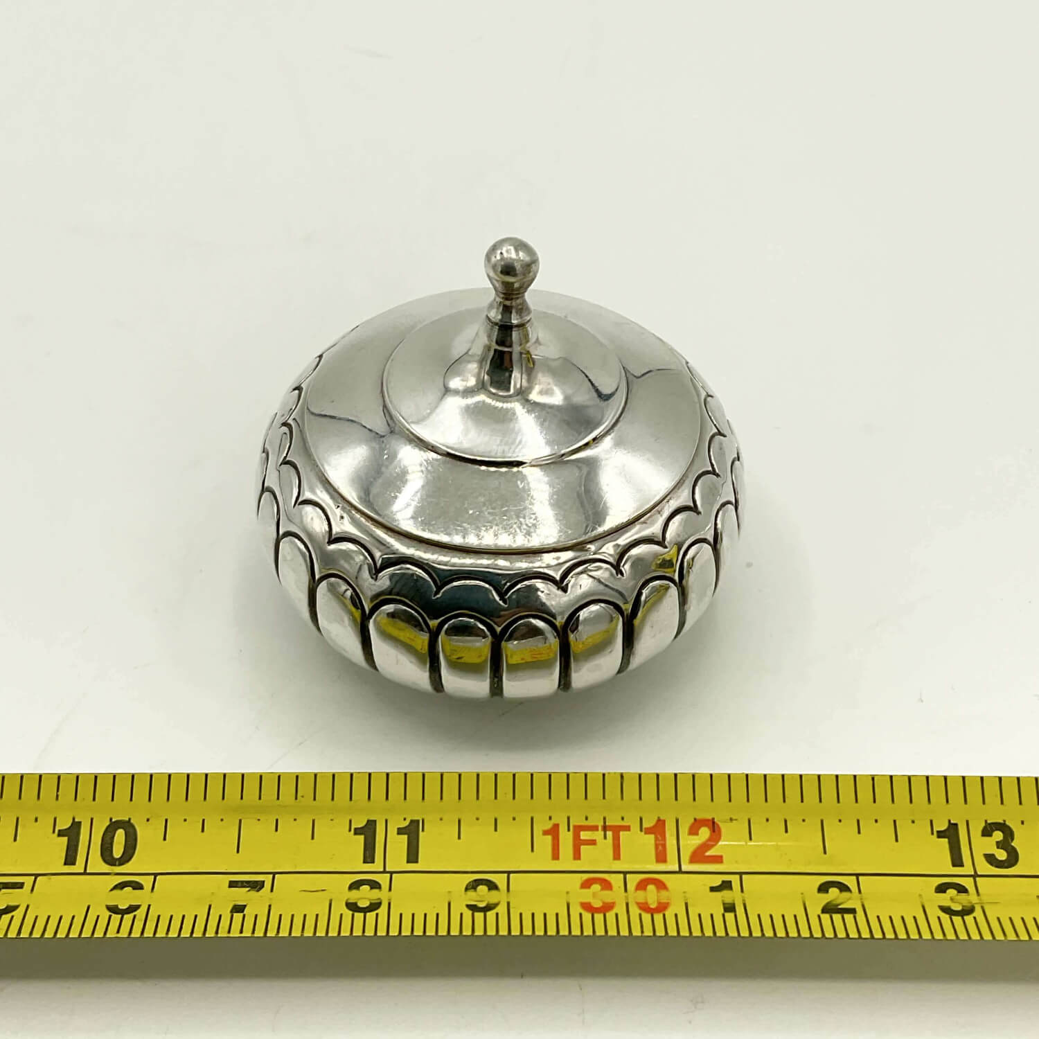 A small round silver box with a lid and decorative scalloped edge, shown on a white background with a yellow measuring tape ruler at the bottom for scale.