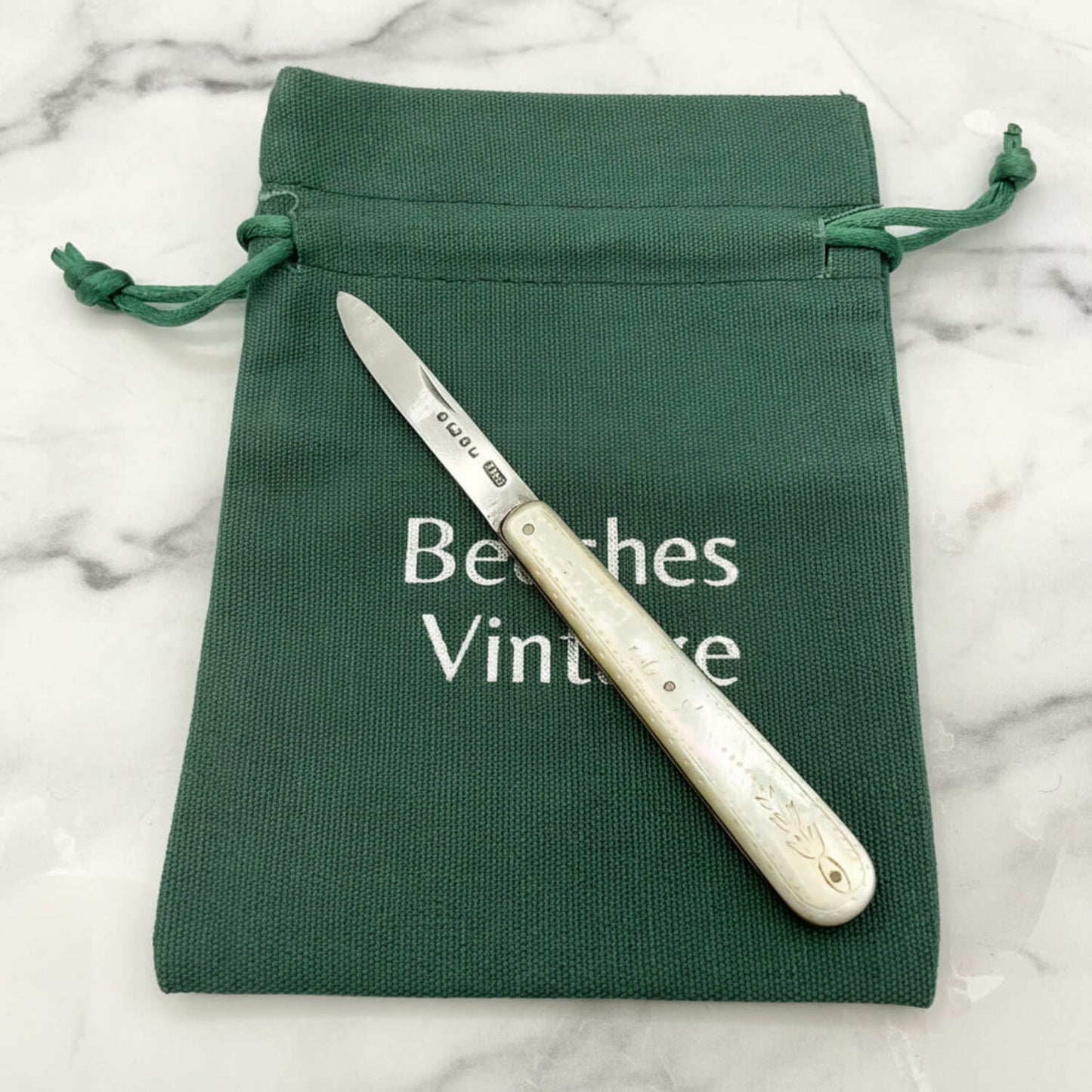 Knife with white handle on a green drawstring bag labeled 'Beeches Vintage' on a marble surface.
