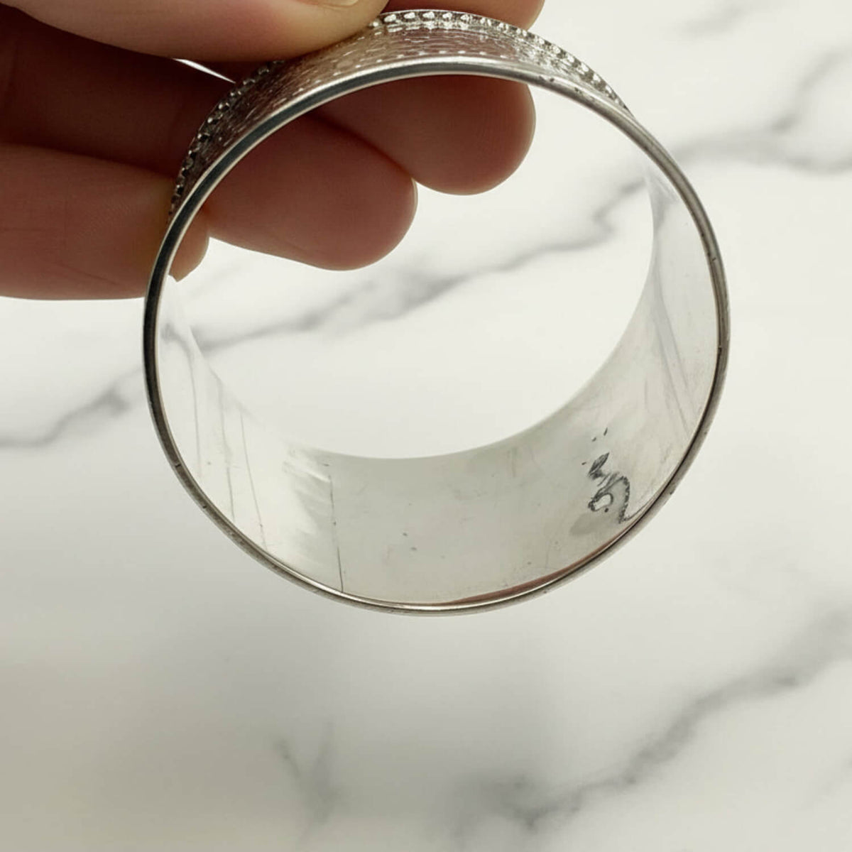 A silver napkin ring with a textured, beaded top band, held by a hand over a white marble background.