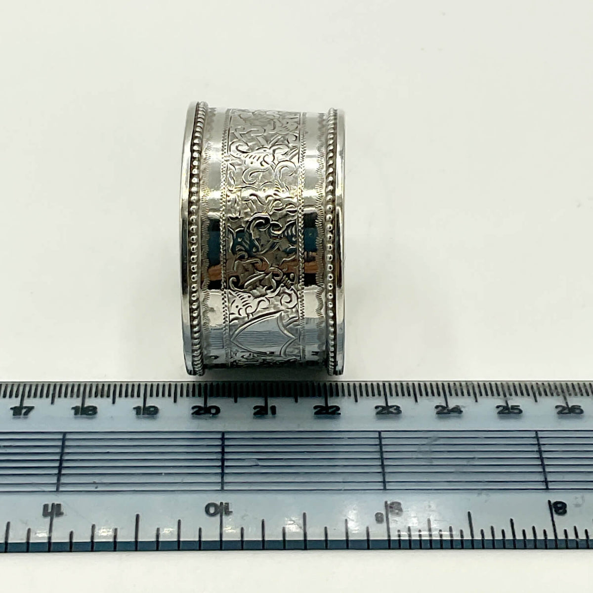 A silver napkin ring with ornate floral and geometric patterns, shown upright next to a ruler for scale on a plain white background.
