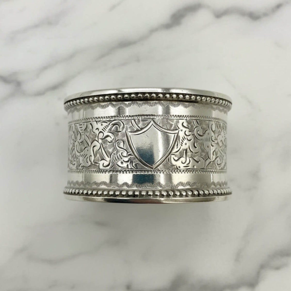 An ornate sterling silver napkin ring intricate floral engravings, a central shield motif, and beaded borders, displayed on a white marble surface.