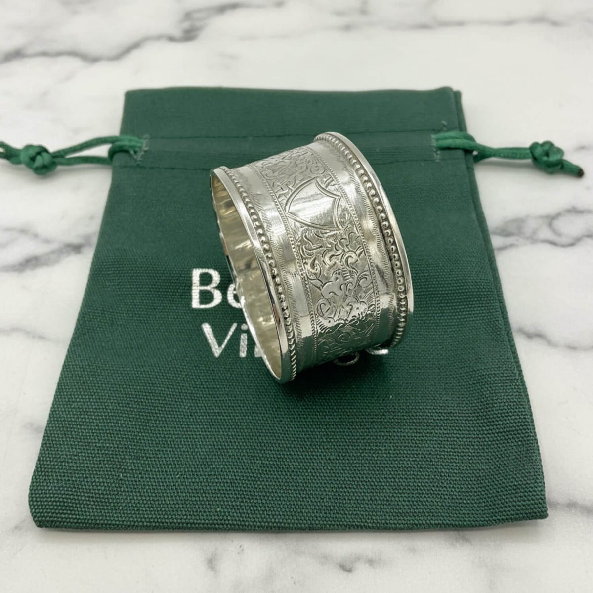 An ornate silver napkin ring with intricate engraved patterns, resting on a dark green drawstring pouch with white text.