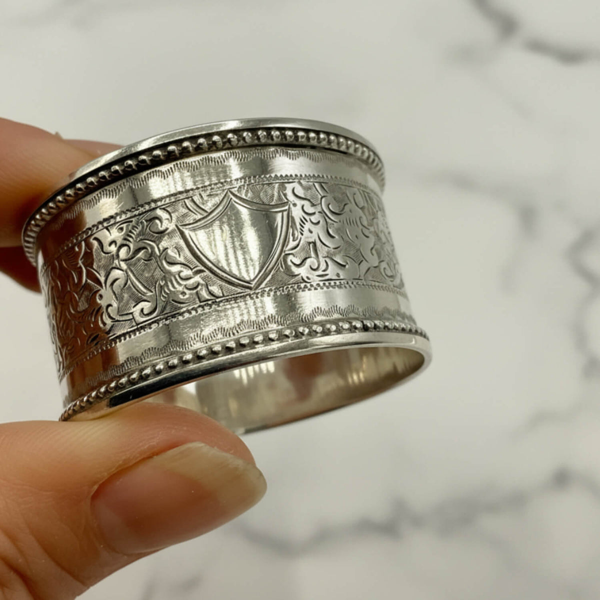 A close-up of a hand holding a wide silver engraved ring with a shield motif and decorative scrollwork, against a plain light background.