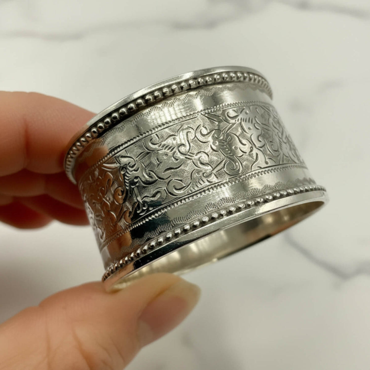 A hand holding a wide silver napkin ring with ornate floral scrollwork and beaded borders, set against a light marble-like background.