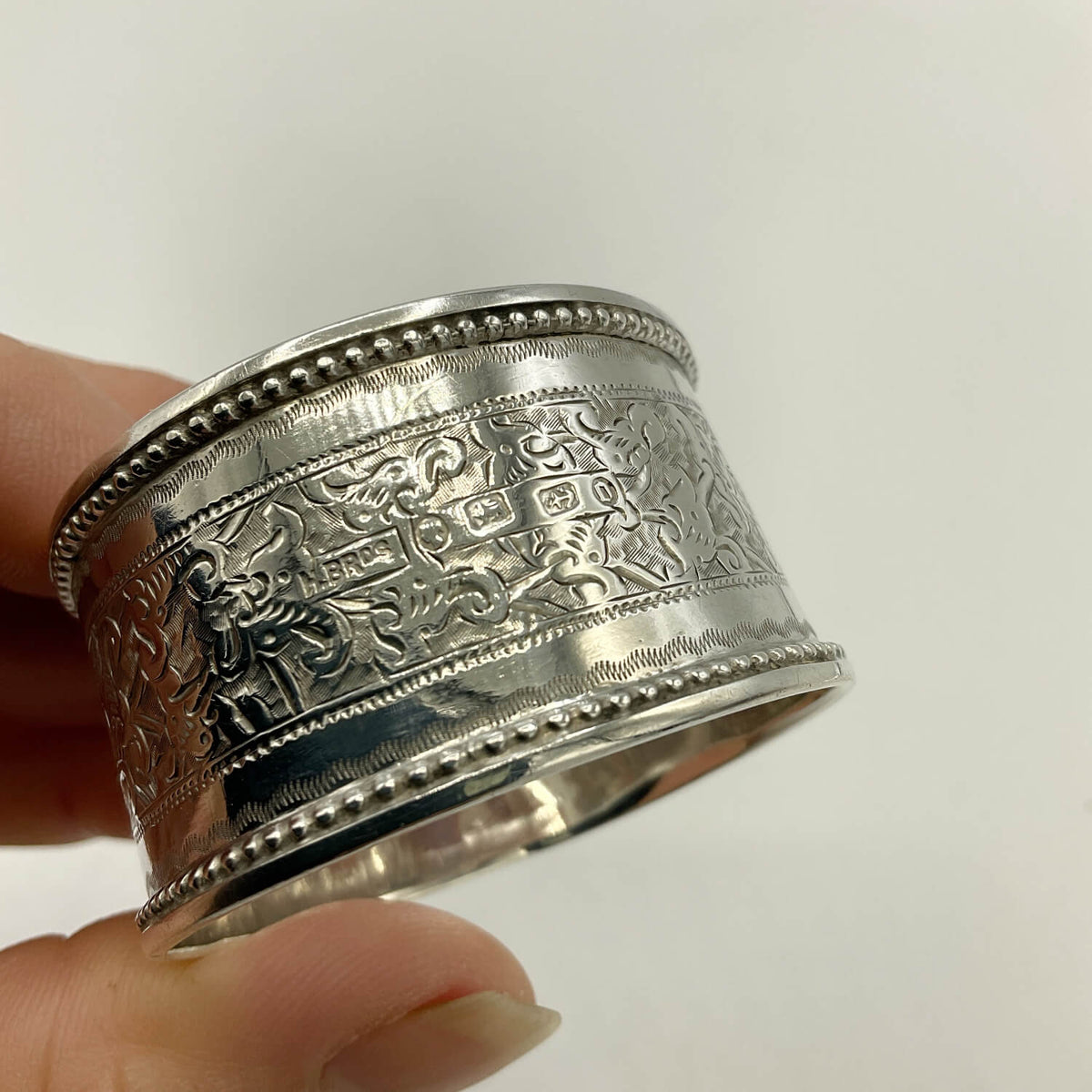 A close-up of a hand holding a wide silver engraved ring with decorative relief patterns and beaded borders, against a plain light background.