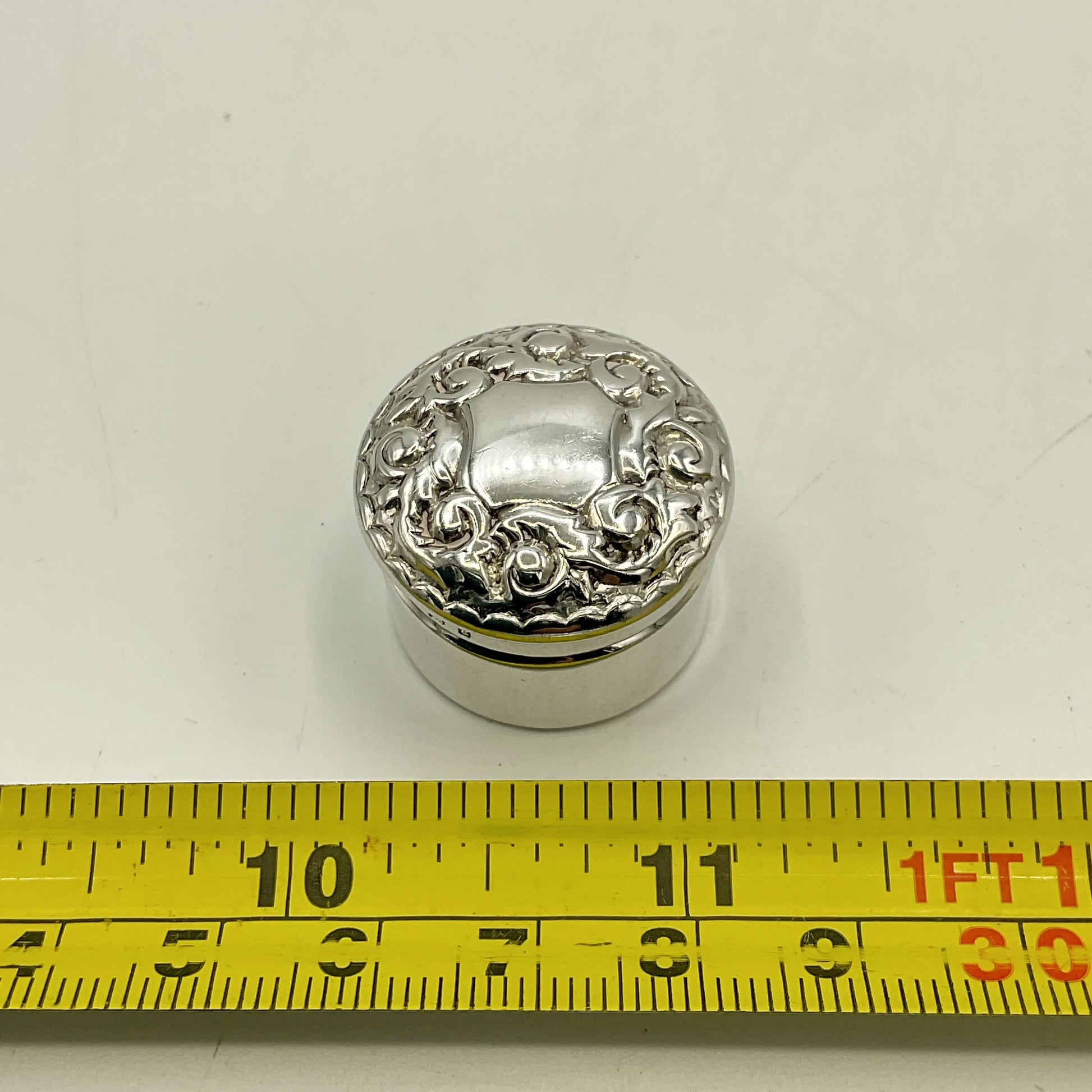 Silver decorative round pill box with intricate patterns on a white background, measured by a yellow tape measure.