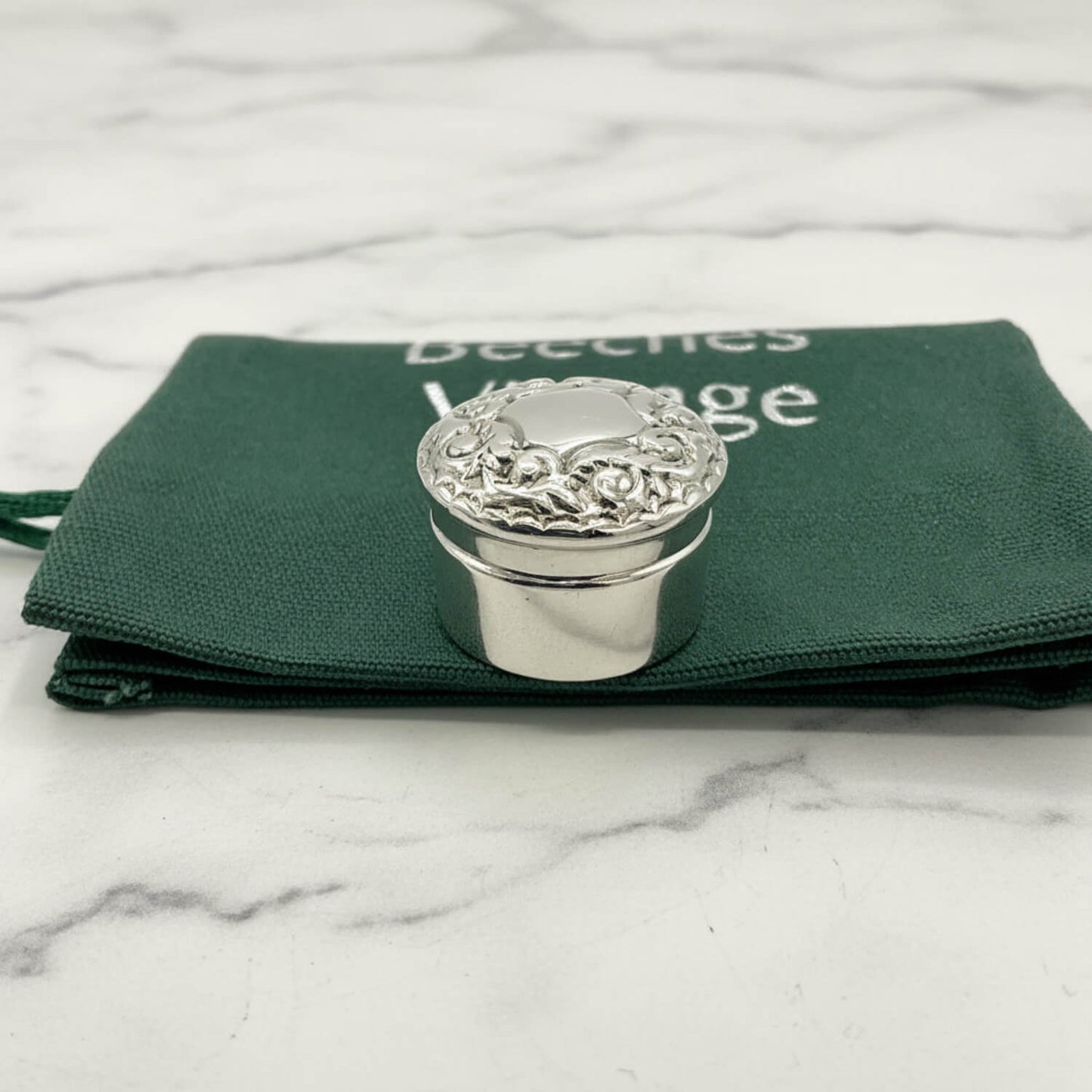 Silver decorative pill box on a green fabric bag with a marble background