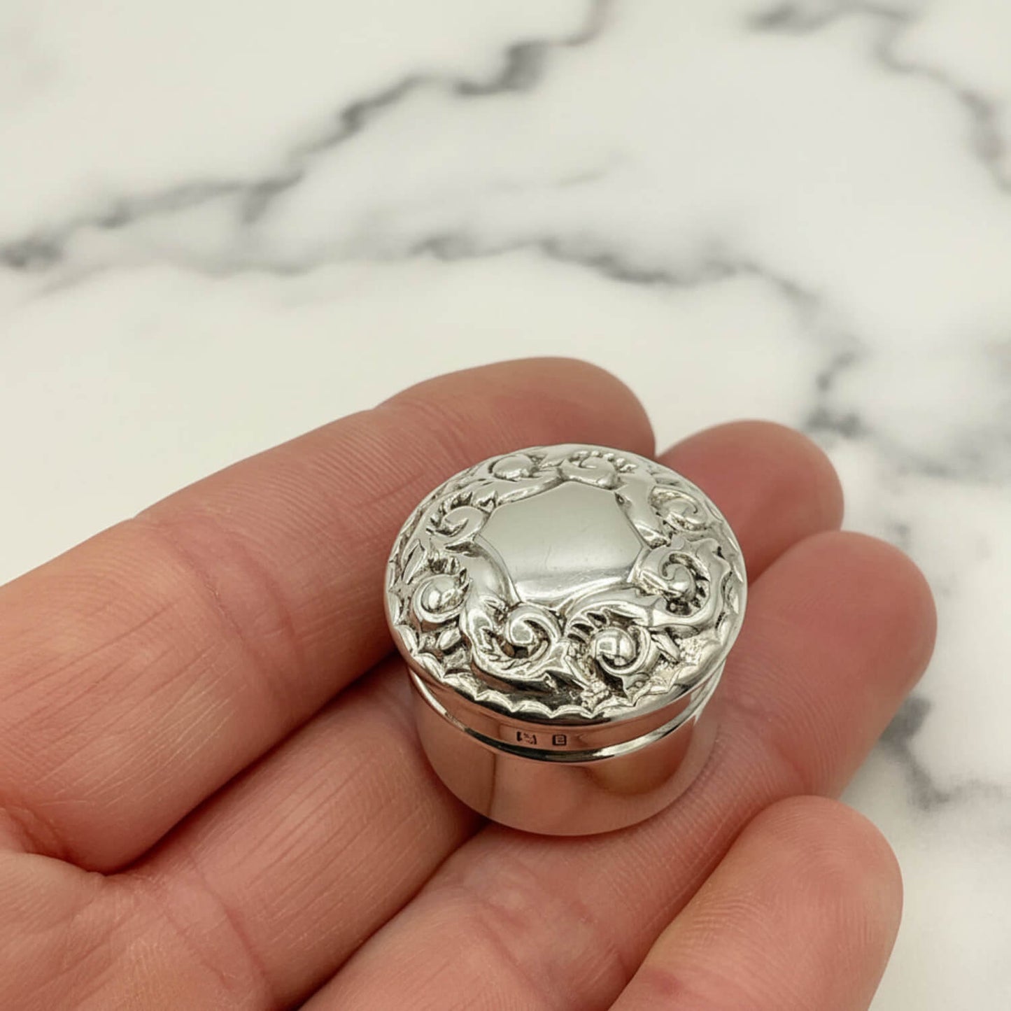 Silver decorative pill box held in a hand with a marble background