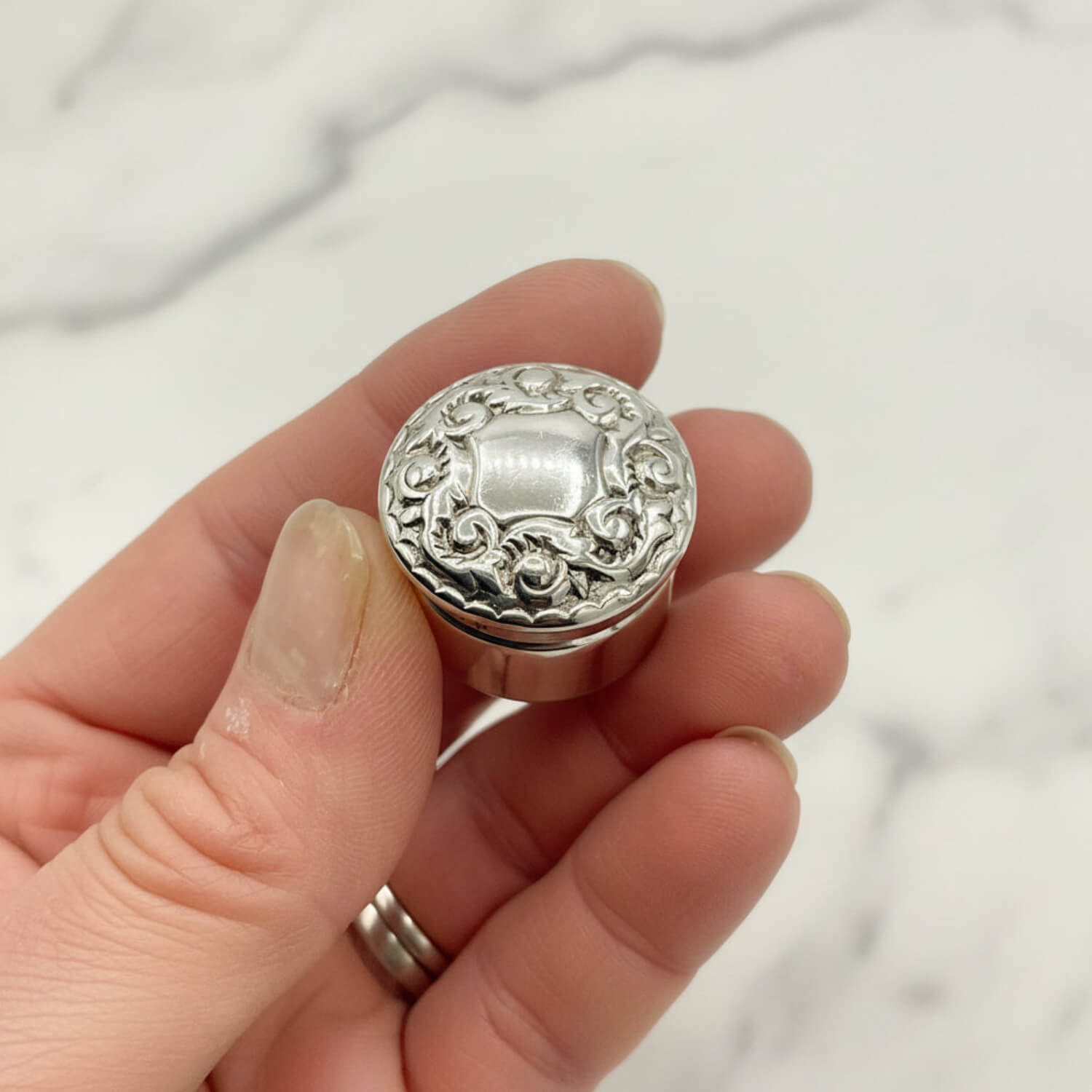 Silver decorative pill box held in a hand against a neutral background