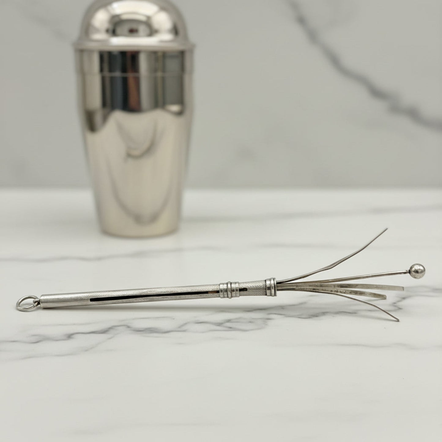 A polished silver cocktail whisk resting on a white marble surface, with a matching silver cocktail shaker blurred in the background.
